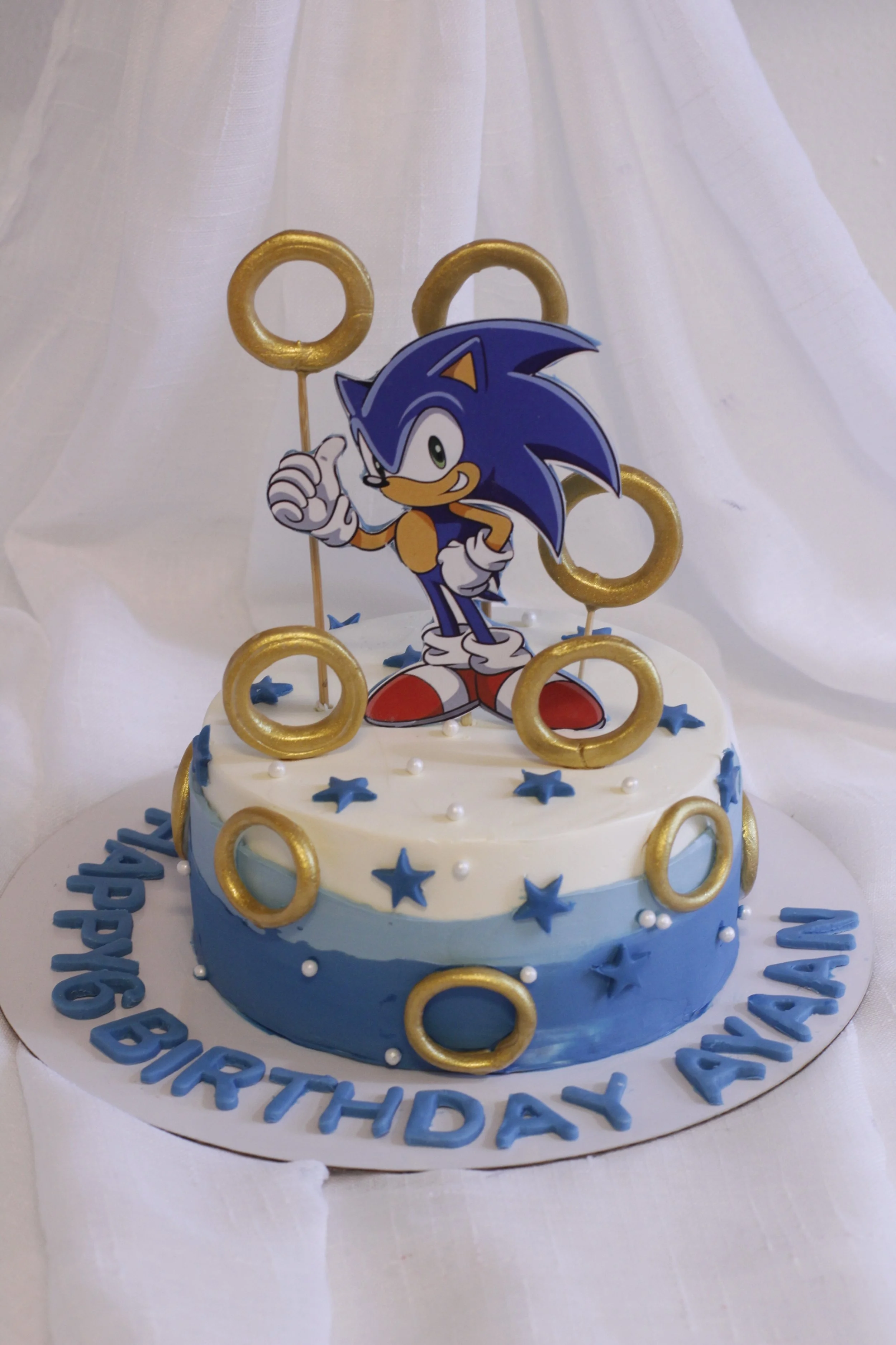 A birthday cake decorated with Sonic the Hedgehog theme, featuring a Sonic character cutout on top, blue and white icing, gold rings, blue stars, and birthday message in blue letters reading "HAPPY BIRTHDAY AVAN" on a white circular cake board.