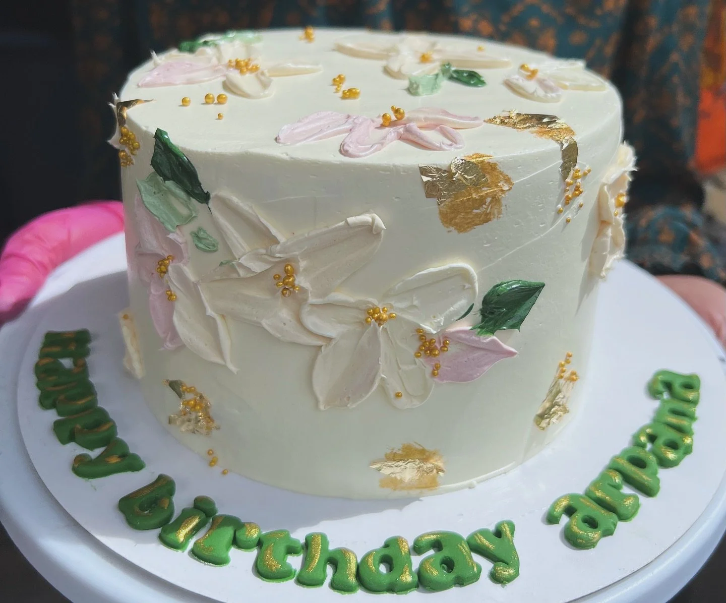 A white birthday cake decorated with pink and white flowers, green leaves, gold accents, and edible gold leaf. The cake sits on a white board with green and gold writing that reads "Happy Birthday".