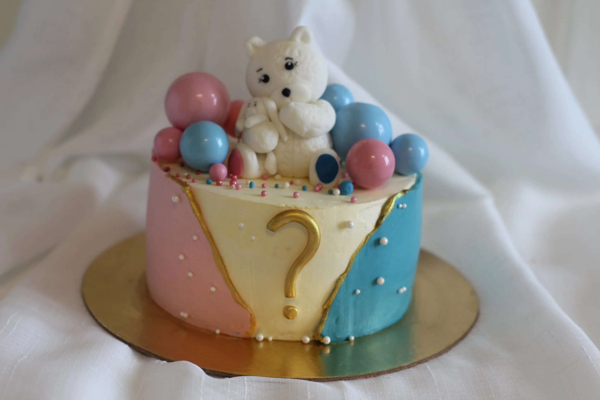 Colorful birthday cake with a white bear figurine on top, decorated with pastel pink, blue, and white spheres, and a gold question mark on the side.