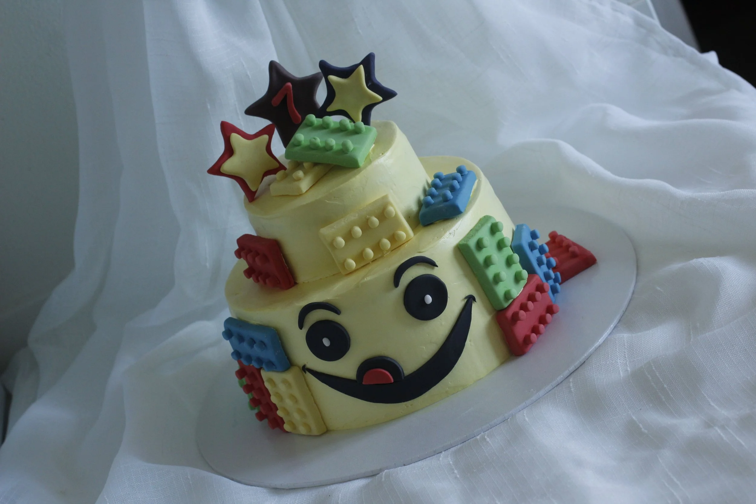 A two-tiered birthday cake decorated with a smiling face, surrounded by colorful building block toppers, on a white cloth background.