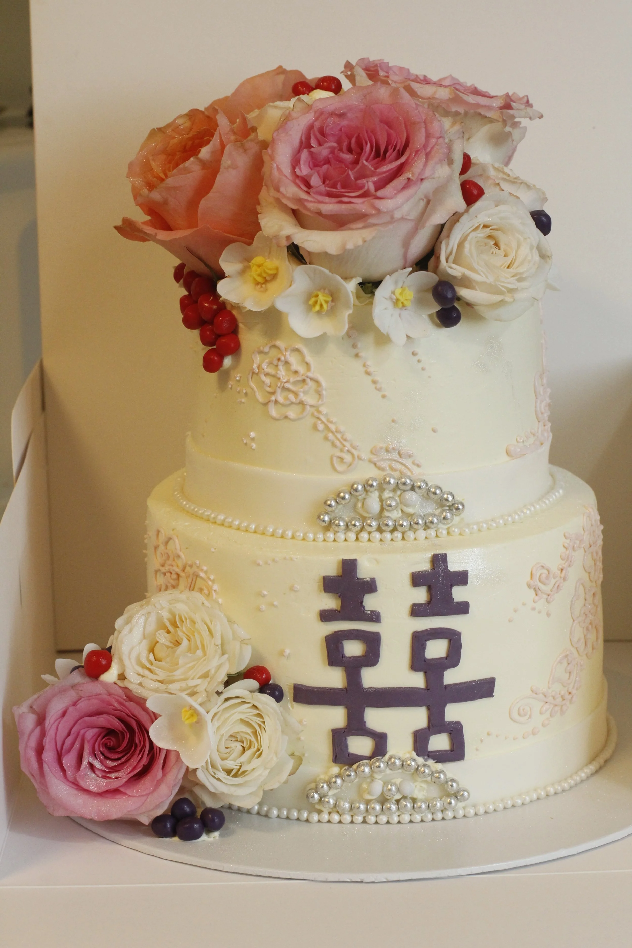 A two-tier wedding cake decorated with pink and white flowers, berries, pearl-like beads, and Chinese characters on the front.