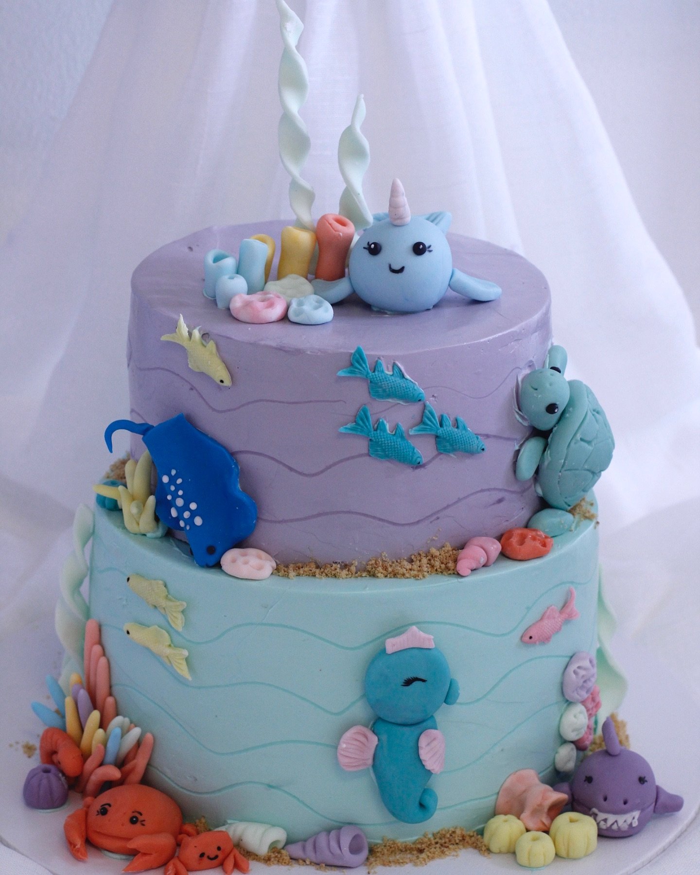 Two-tiered cake decorated with pastel-colored fondant ocean creatures like a unicorn, seahorses, fish, a whale, a turtle, and seaweed, with a white tulle fabric background.