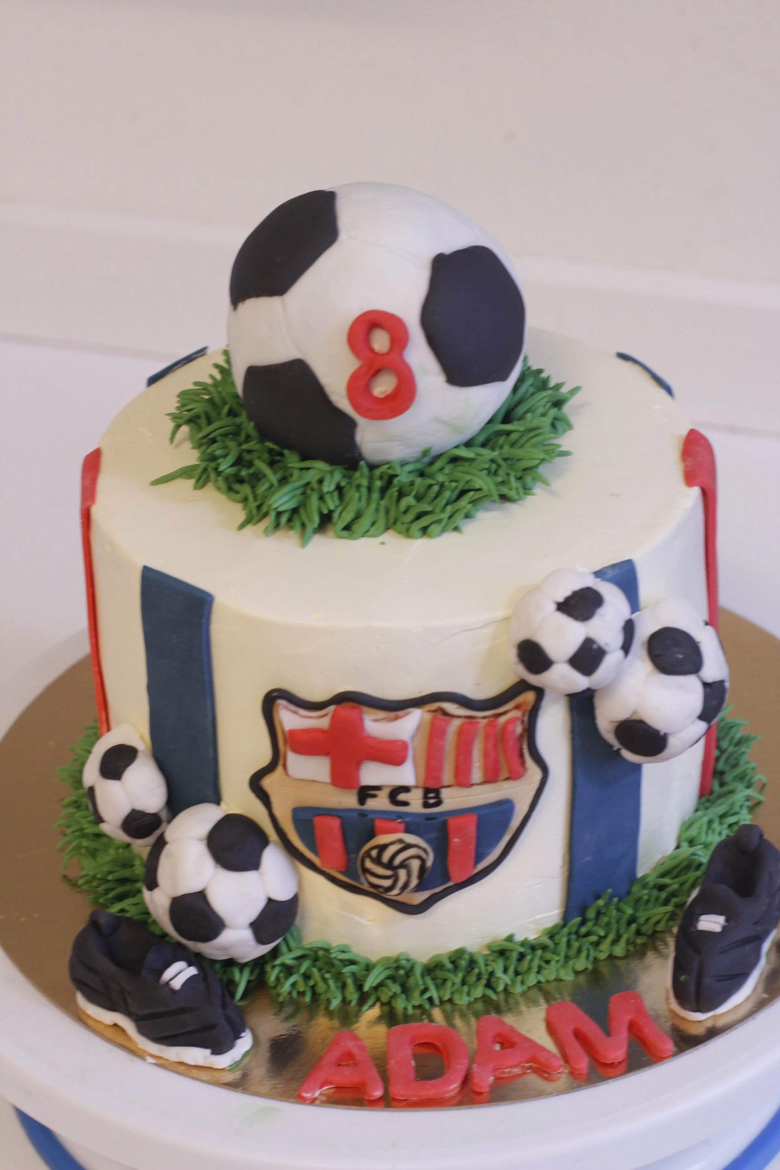 A soccer-themed birthday cake decorated with black and white soccer balls, a FC Barcelona crest, and a soccer ball with the number 8 on top. The cake has green icing around the base and the name "ADAM" in red letters.