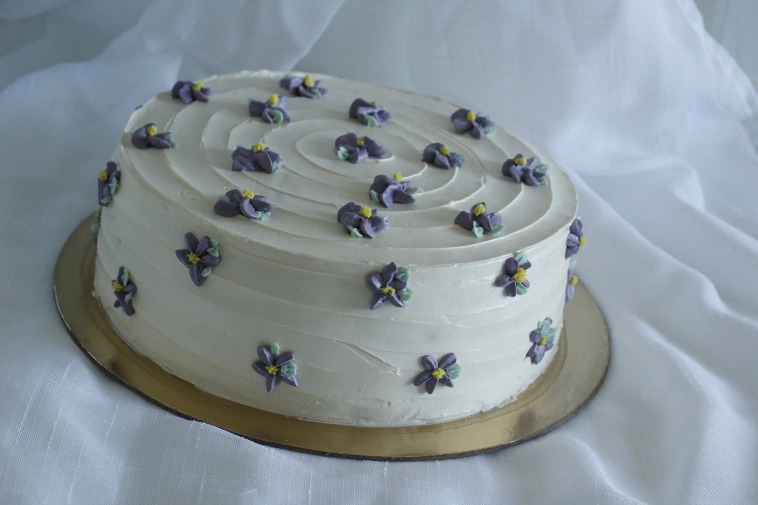 A heart-shaped white wedding cake decorated with small purple and yellow flower decorations on top and around the sides, placed on a gold cake board on white fabric.