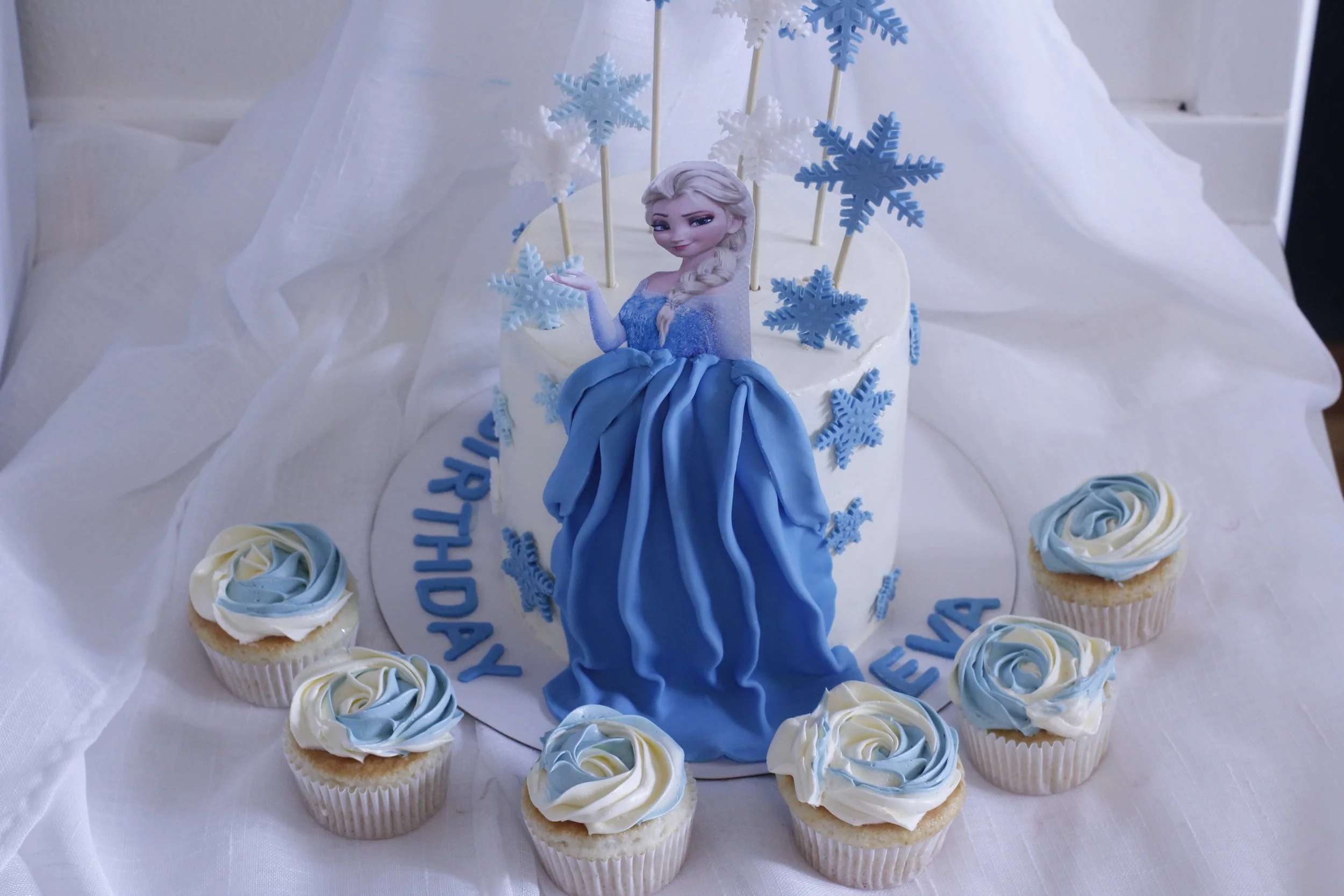 Frozen themed birthday cake with an image of Elsa and blue and white snowflake decorations, surrounded by cupcakes with blue and white swirl icing.