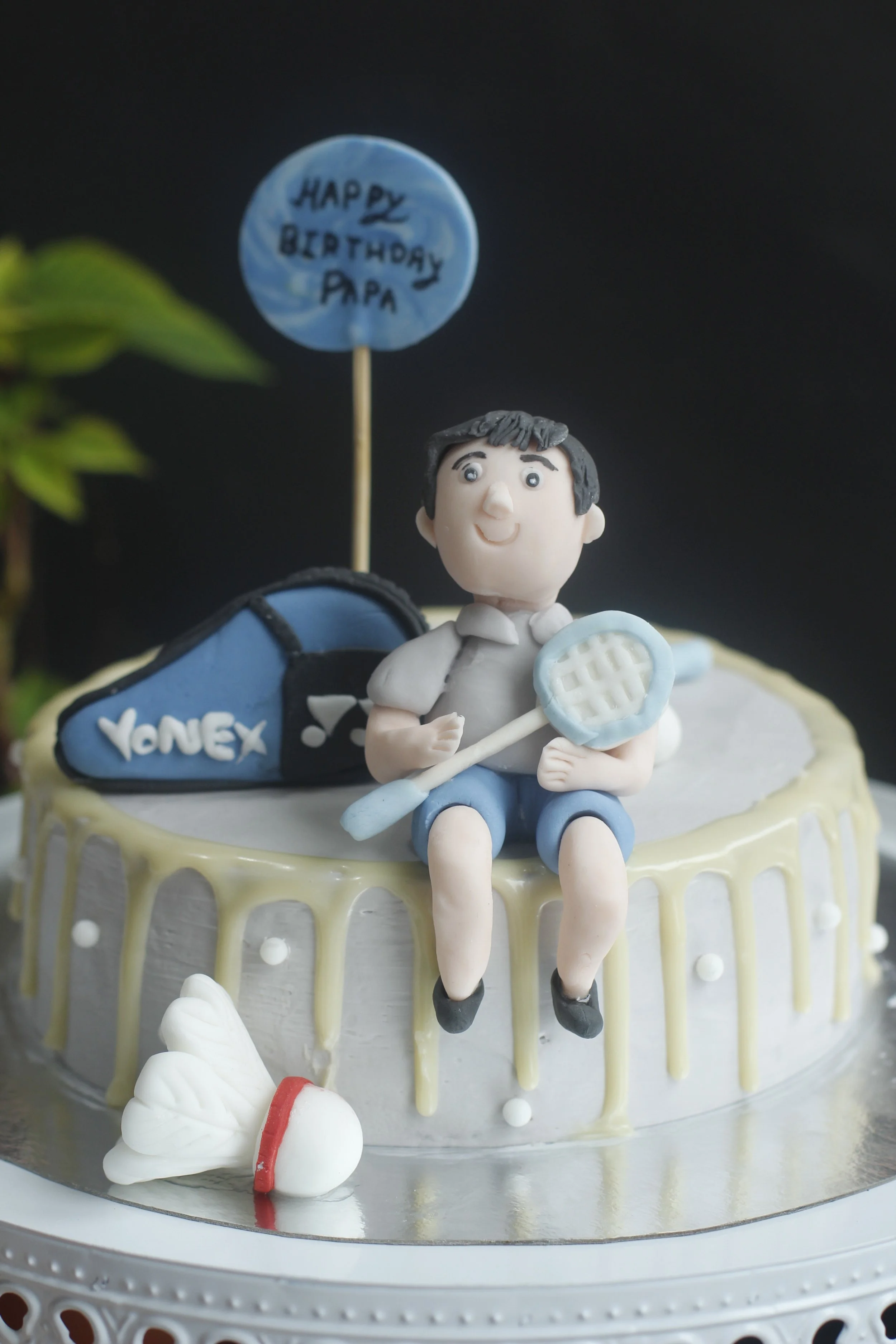 A birthday cake with a tennis-themed topper, featuring a fondant figure of a boy holding a tennis racket. There is a tennis bag with 'YONEX' written on it, a sign that reads 'HAPPY BIRTHDAY PAPA,' and a small tennis ball on the cake.