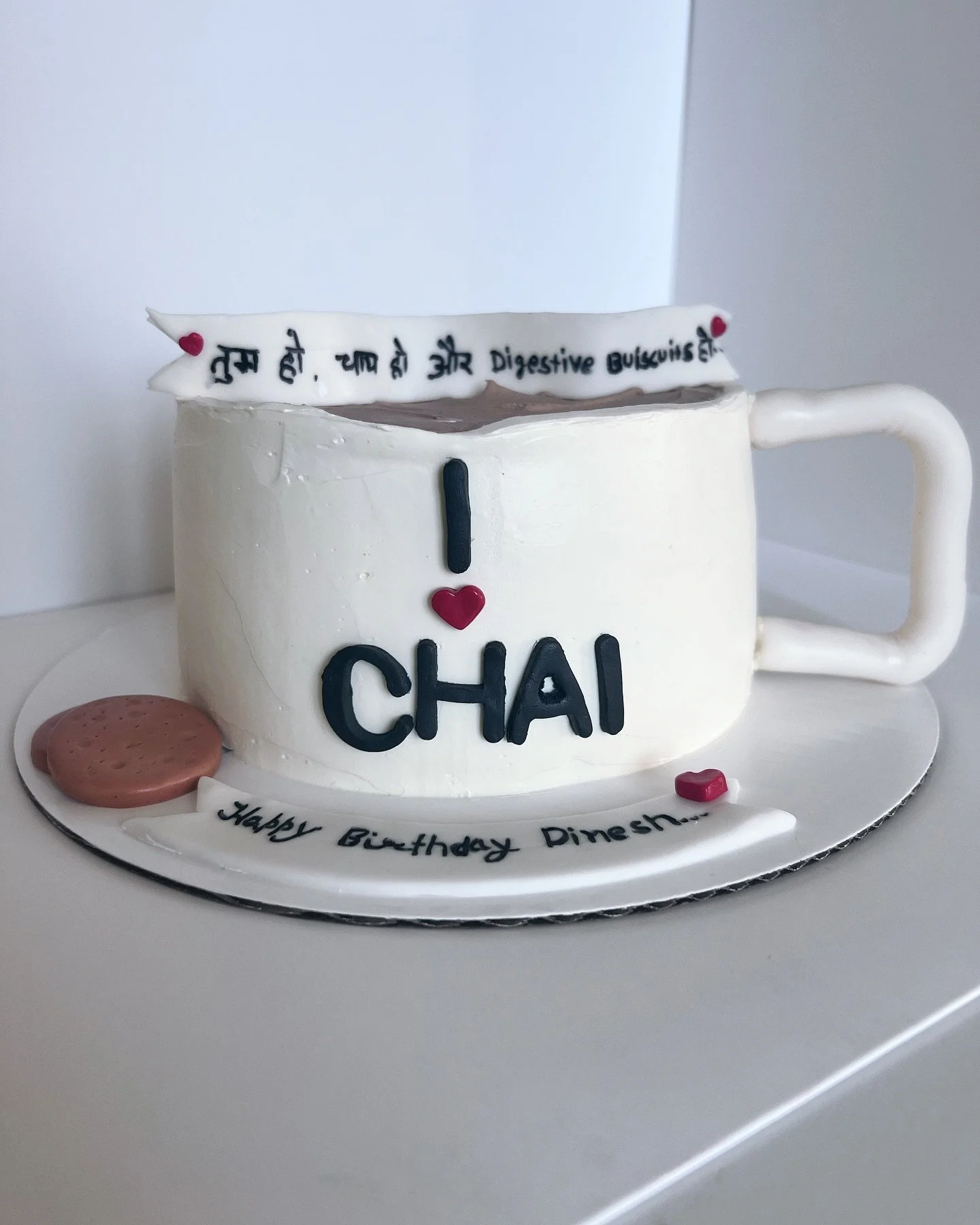 A birthday cake in the shape of a white coffee cup with a handle, decorated with message and heart decorations. The cake has black and red lettering that says "I love Chai" and "Happy Birthday Dinesha". There are some small decorations resembling coo