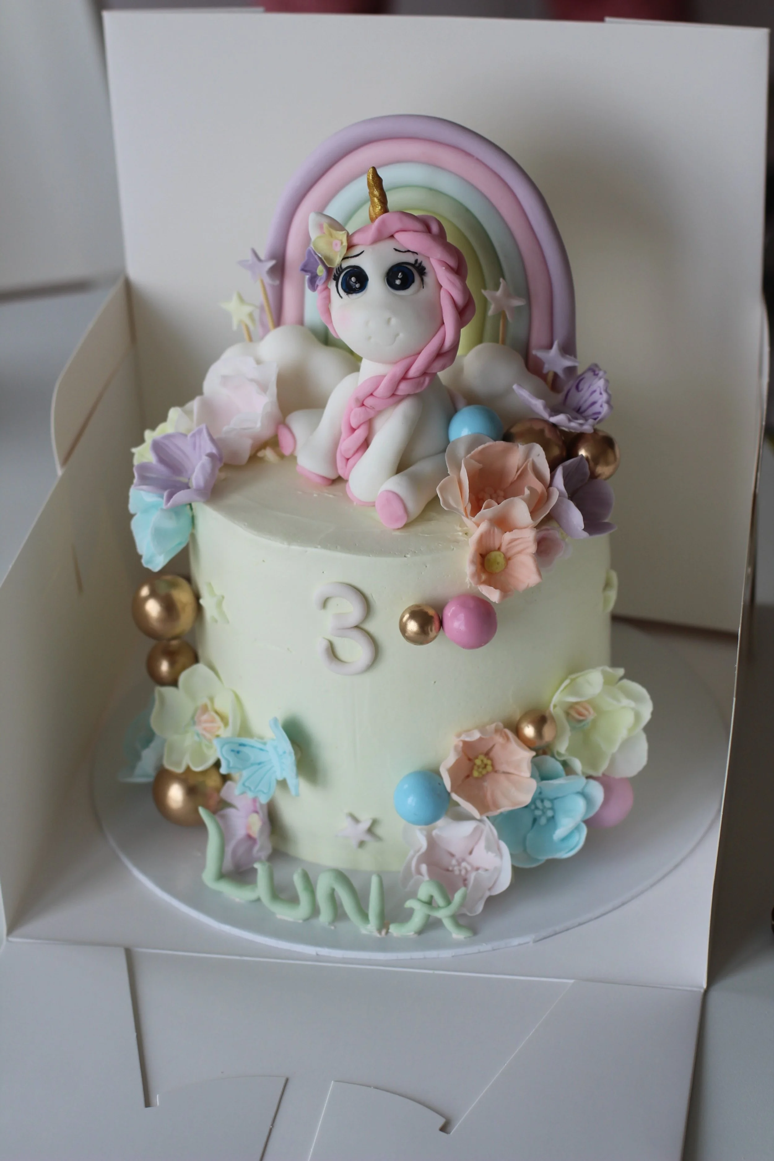 A pastel-colored birthday cake featuring a unicorn figure with a pink mane and rainbow horn, decorated with flowers, stars, and balloons, with the number three and the name "Luna" on it.