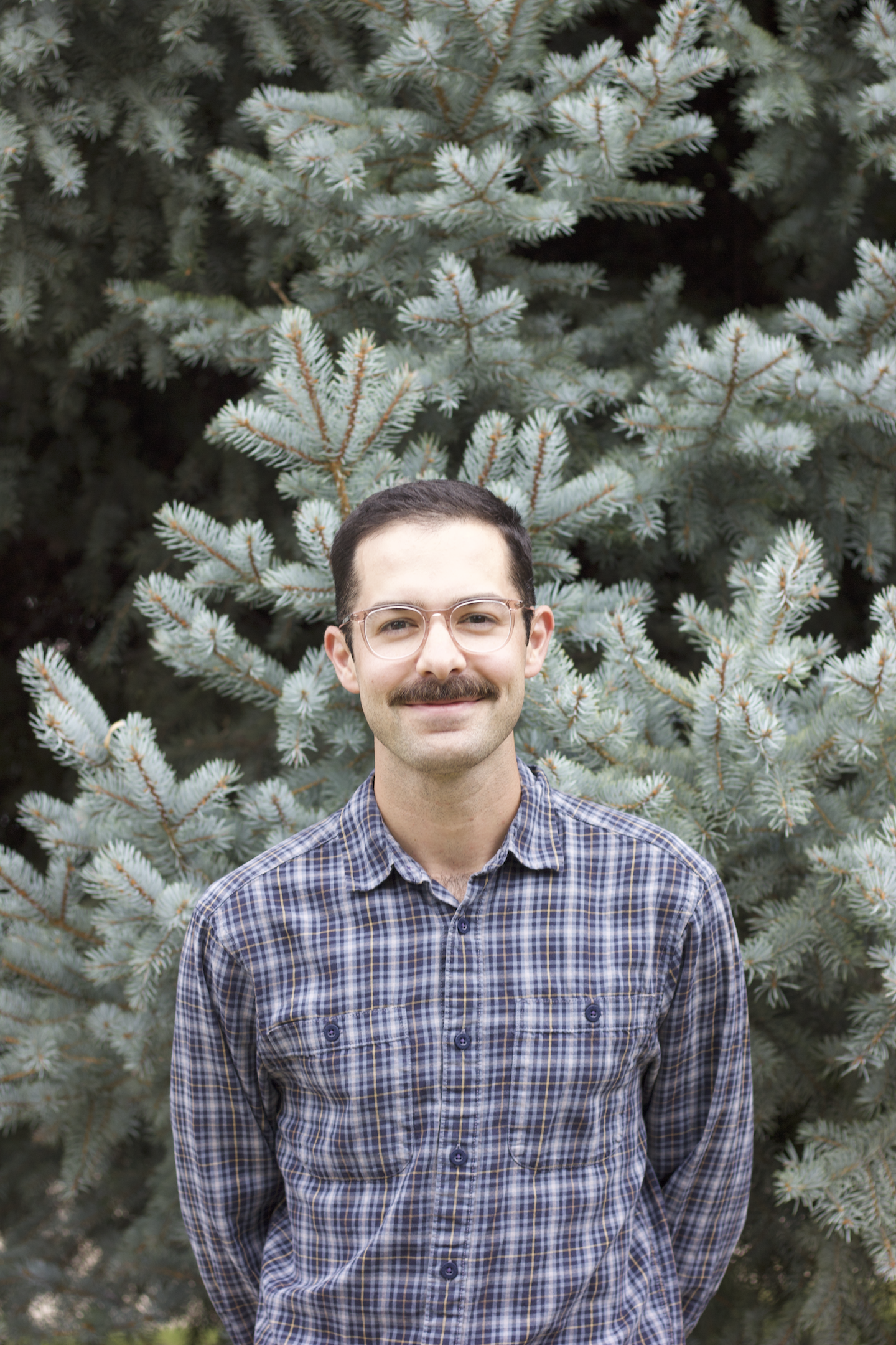 A man with glasses, a mustache, and a plaid shirt standing outdoors in front of a large pine tree.