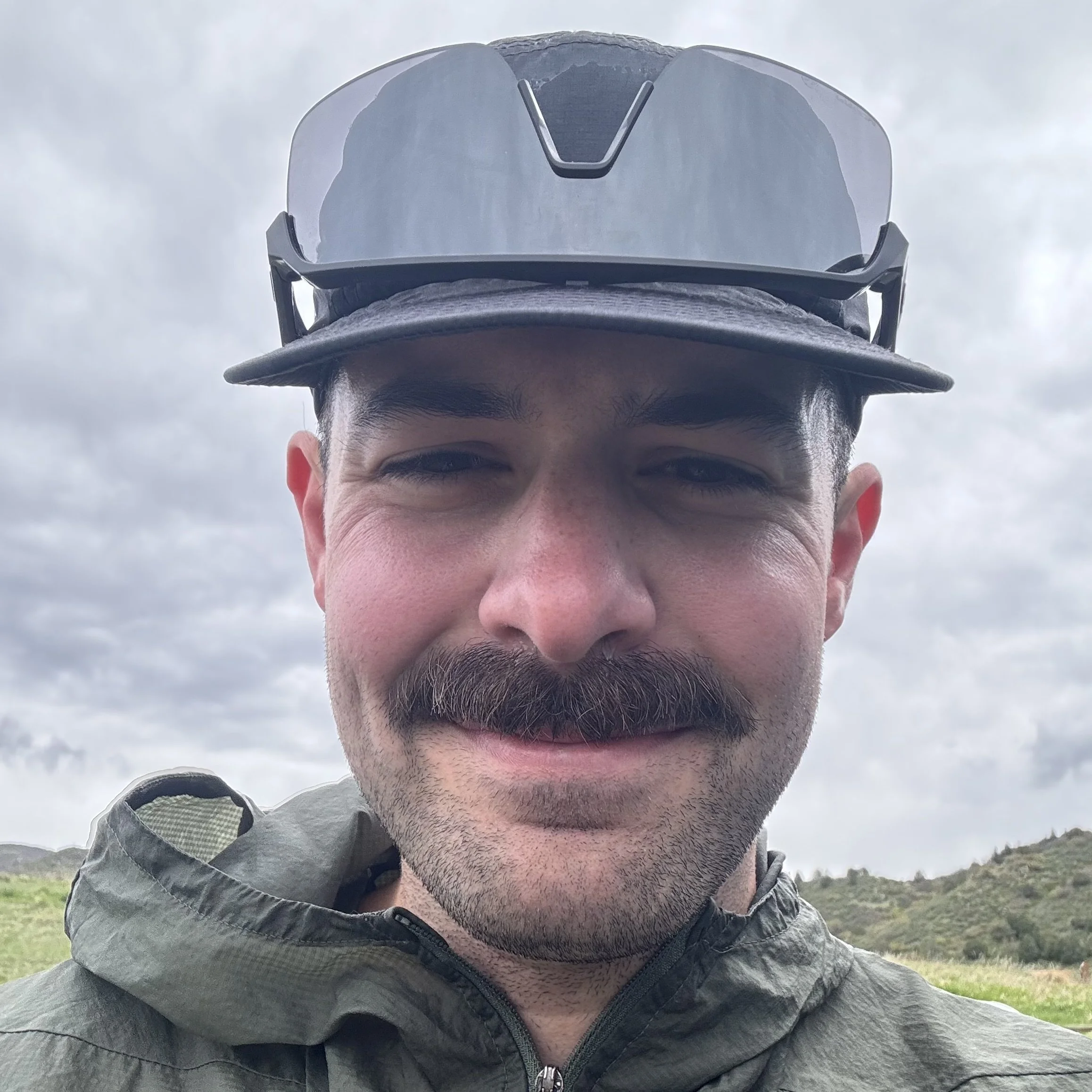A man with a mustache and facial hair outdoors, wearing a gray jacket and sunglasses on his hat, with a cloudy sky and green hills in the background.