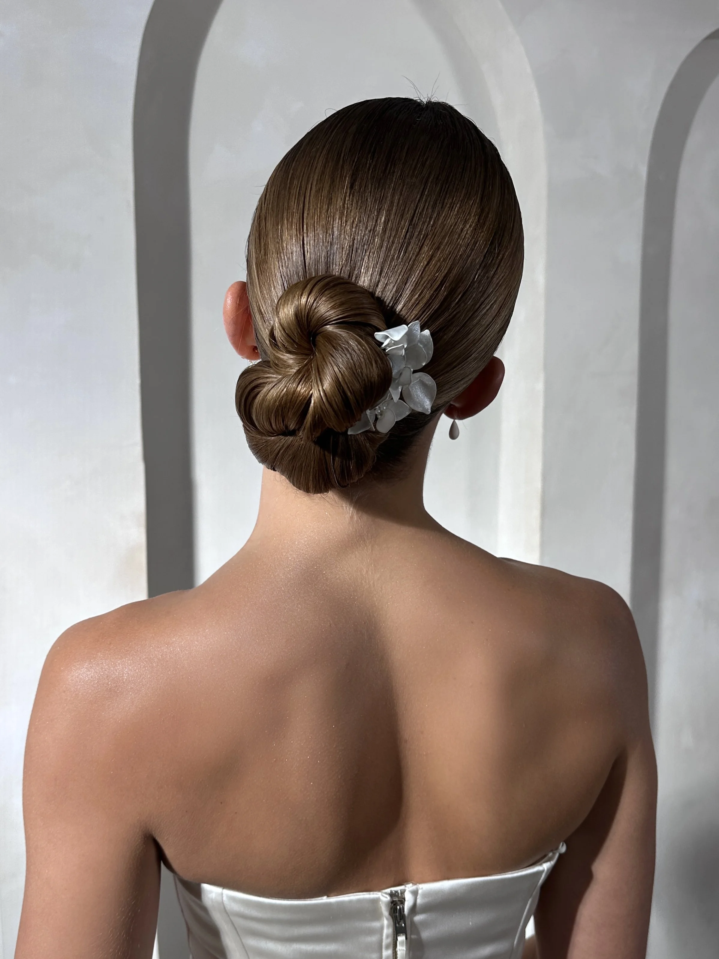 Back view of a woman with smooth, brown hair styled in an elegant bun adorned with white flowers, wearing a strapless white dress with a zipper at the back, against a plain white wall background.