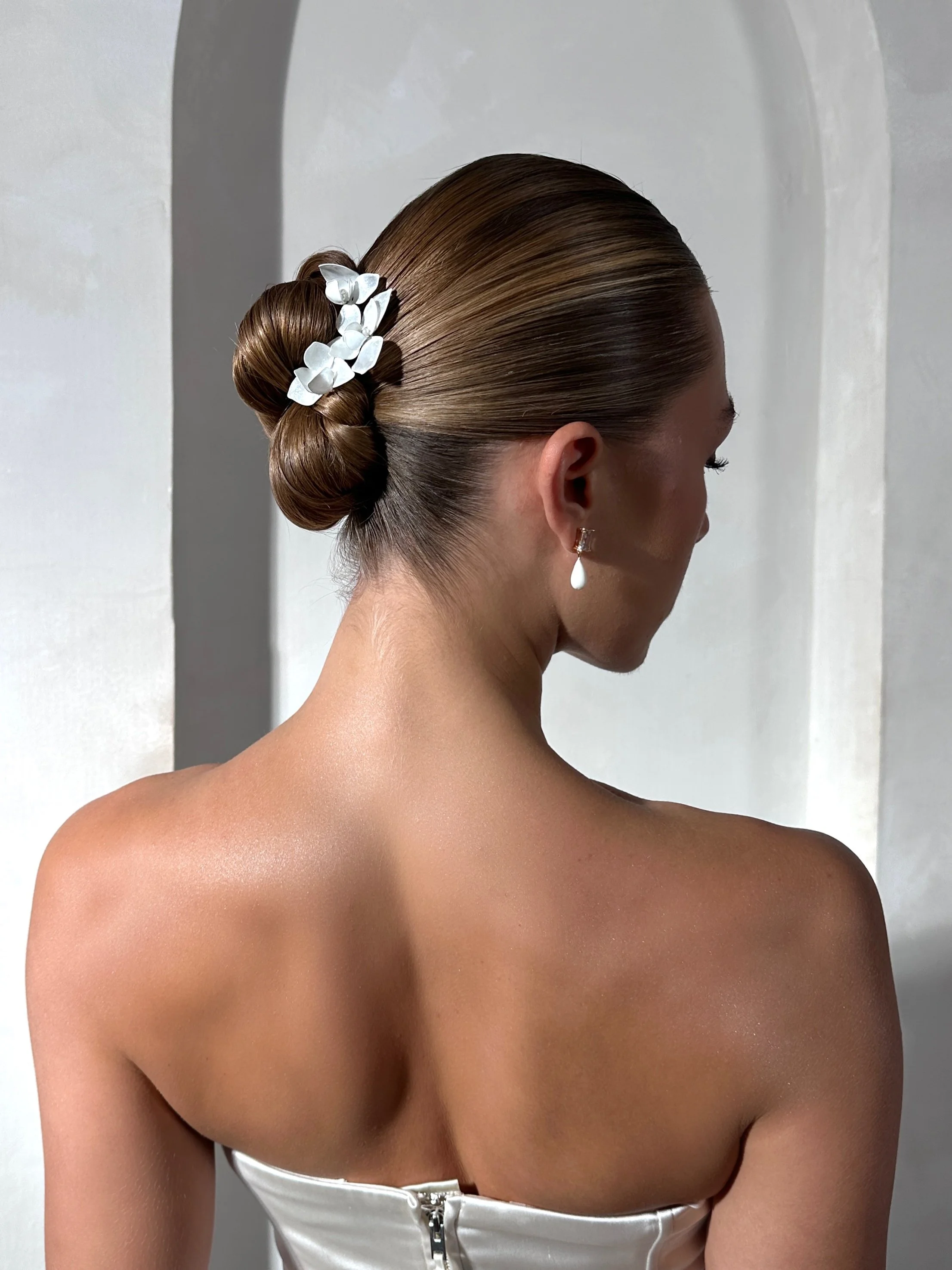 Elegant woman with slicked-back hairstyle adorned with white flowers, wearing pearl earrings and a strapless white dress, standing in front of a white wall.