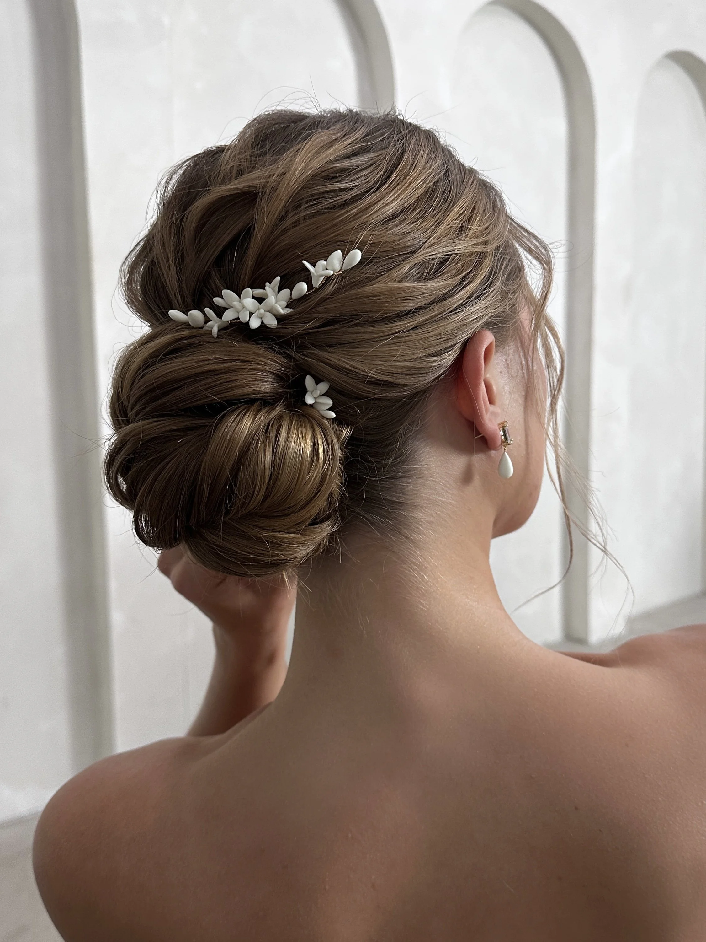 A woman with styled hair adorned with white floral hair accessories, wearing a white gemstone earring, with bare shoulders, facing away from the camera.