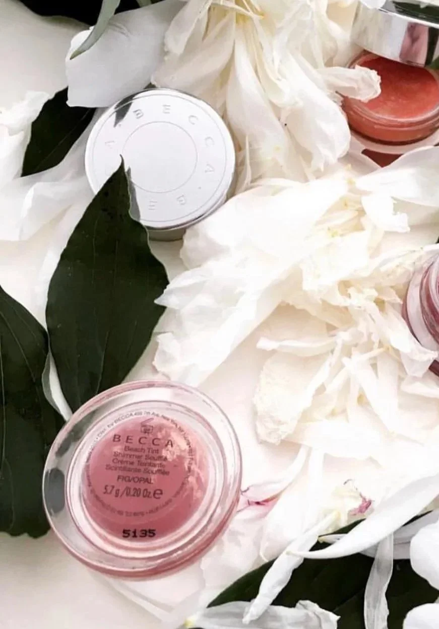 A collection of beauty products and white flowers, including containers labeled with 'BECCA' and 'FOVORI,' displayed among green leaves and white petals.