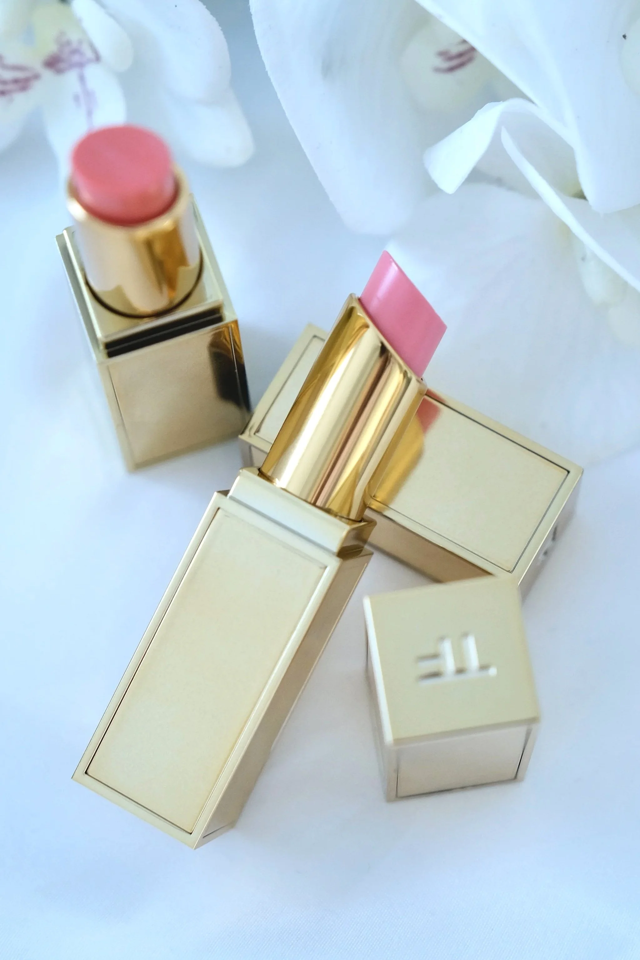 Two lipsticks with gold packaging, one in light pink and the other in a darker pink, placed on white surface with white flowers in background.