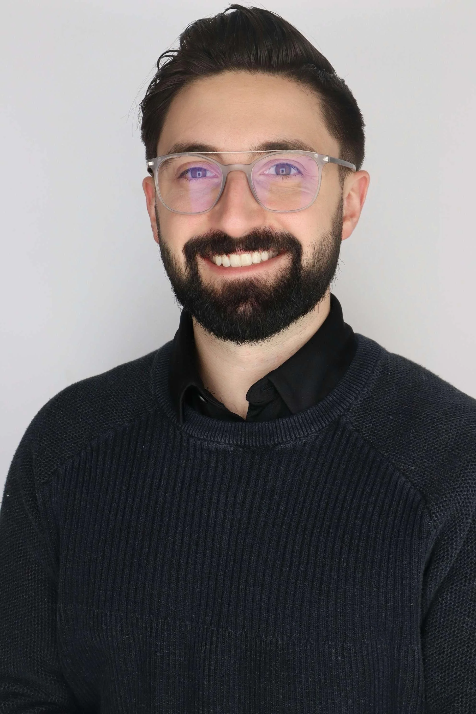 A smiling man with dark hair, glasses with purple-tinted lenses, a beard, and dressed in a black sweater over a black collared shirt, standing against a plain white background.