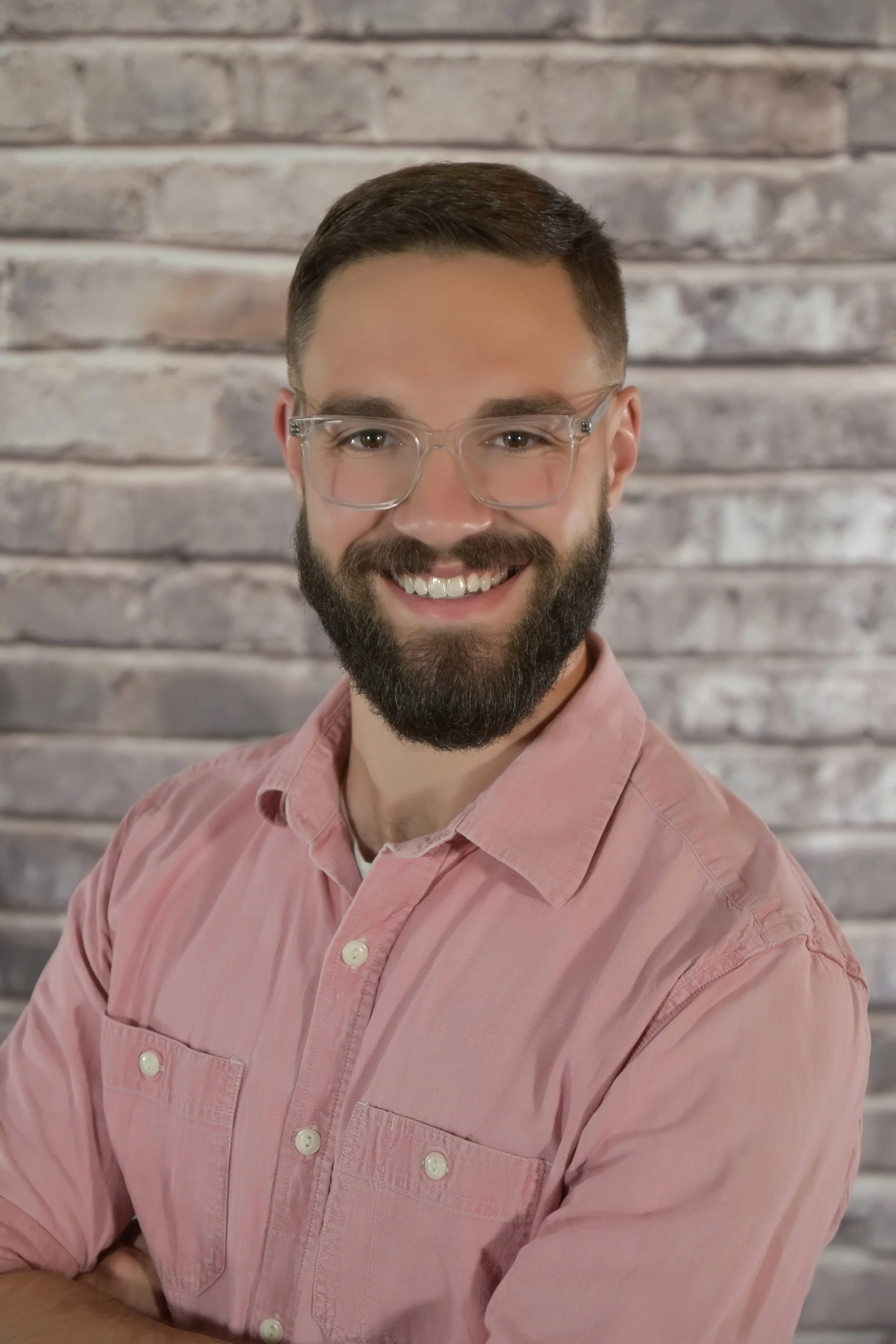 A smiling man with a beard and glasses, wearing a pink button-up shirt, standing in front of a brick wall.