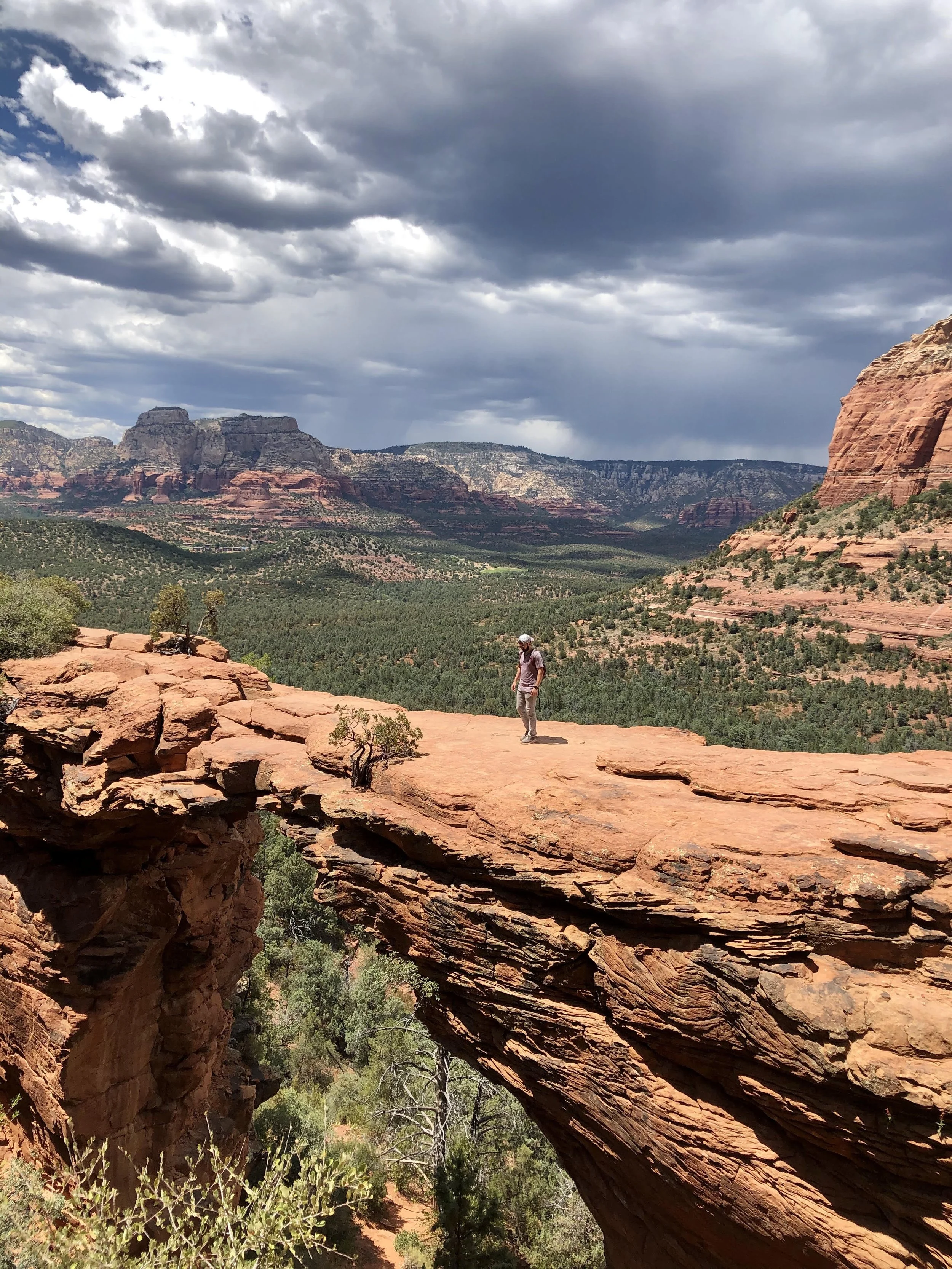 A person standing on a natural rock bridge in a desert landscape with red rocks, green trees, and a cloudy sky.