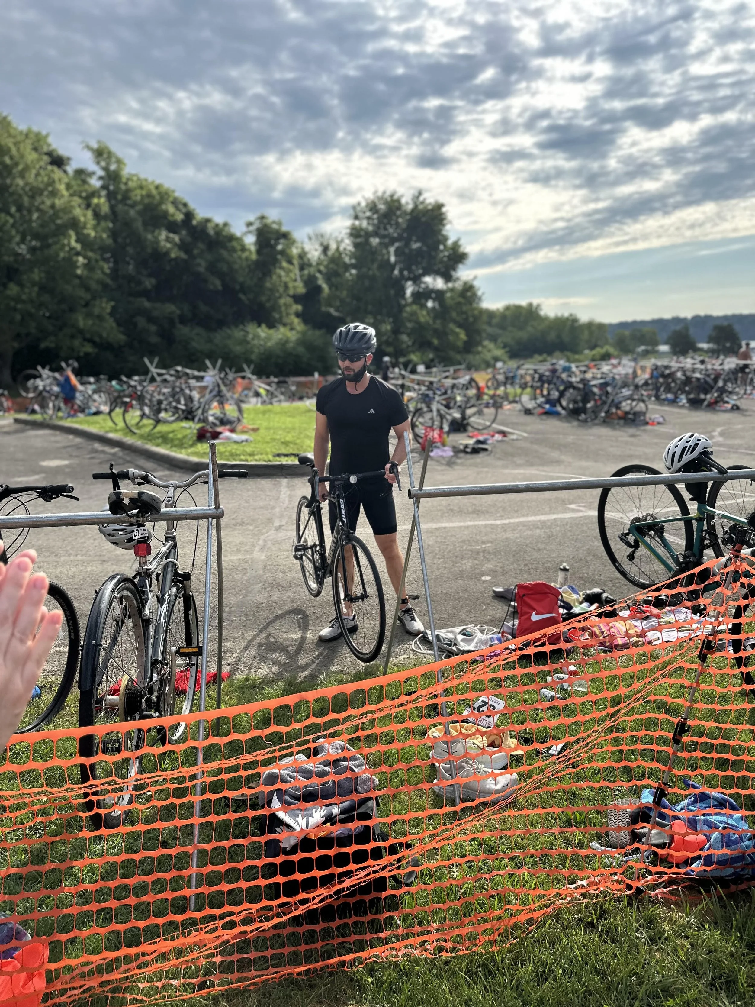 A man in black cycling gear and helmet arranging a bike at a transition area covered with bikes and gear, with many other cyclists in the background and a cloudy sky overhead.