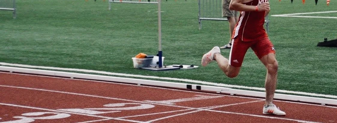 A person running on a track, wearing red athletic clothing and white running shoes, near a running lane marker and a pole with a timer.