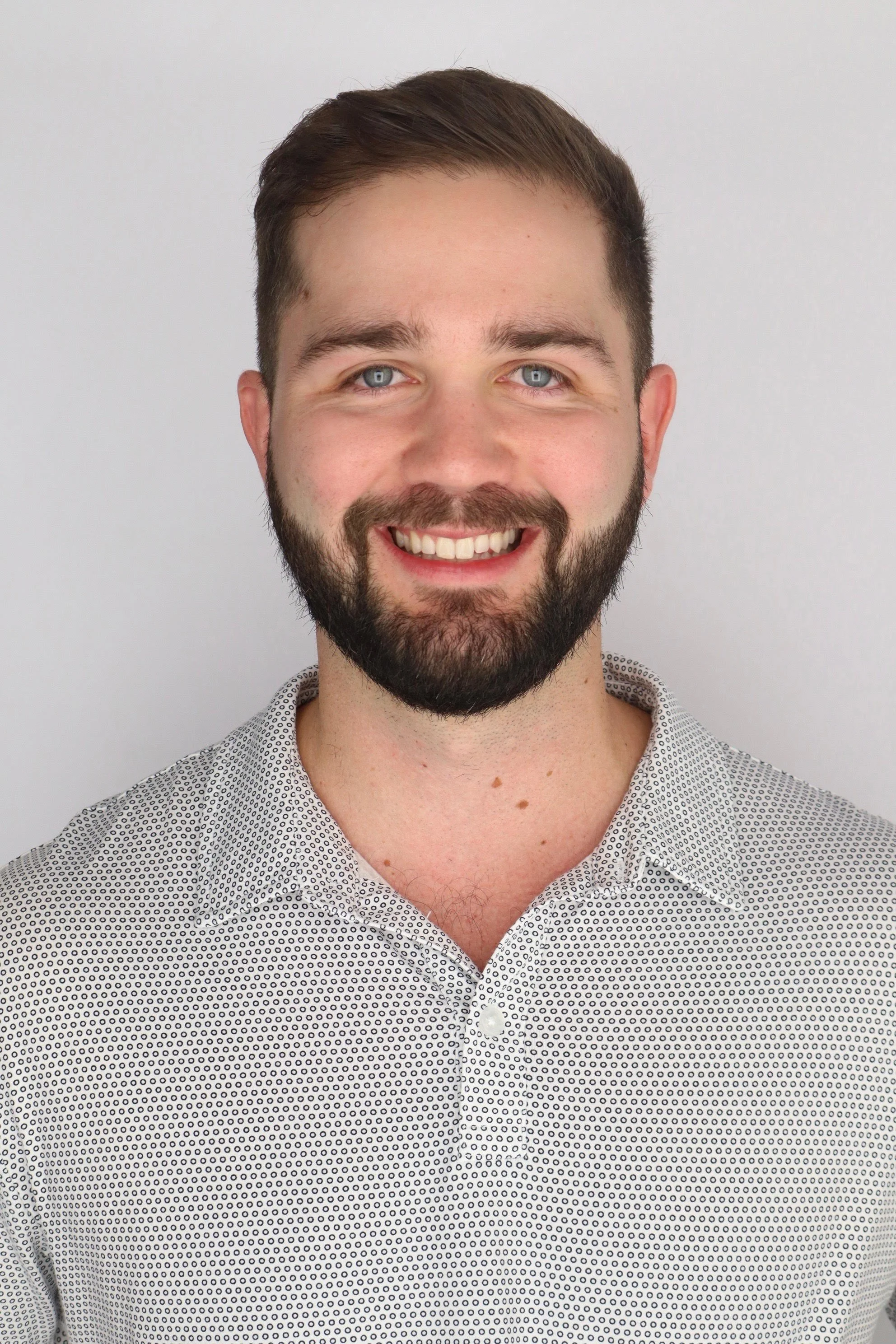 Portrait of a smiling man with a beard and blue eyes, wearing a light-colored patterned shirt, standing against a plain light background.
