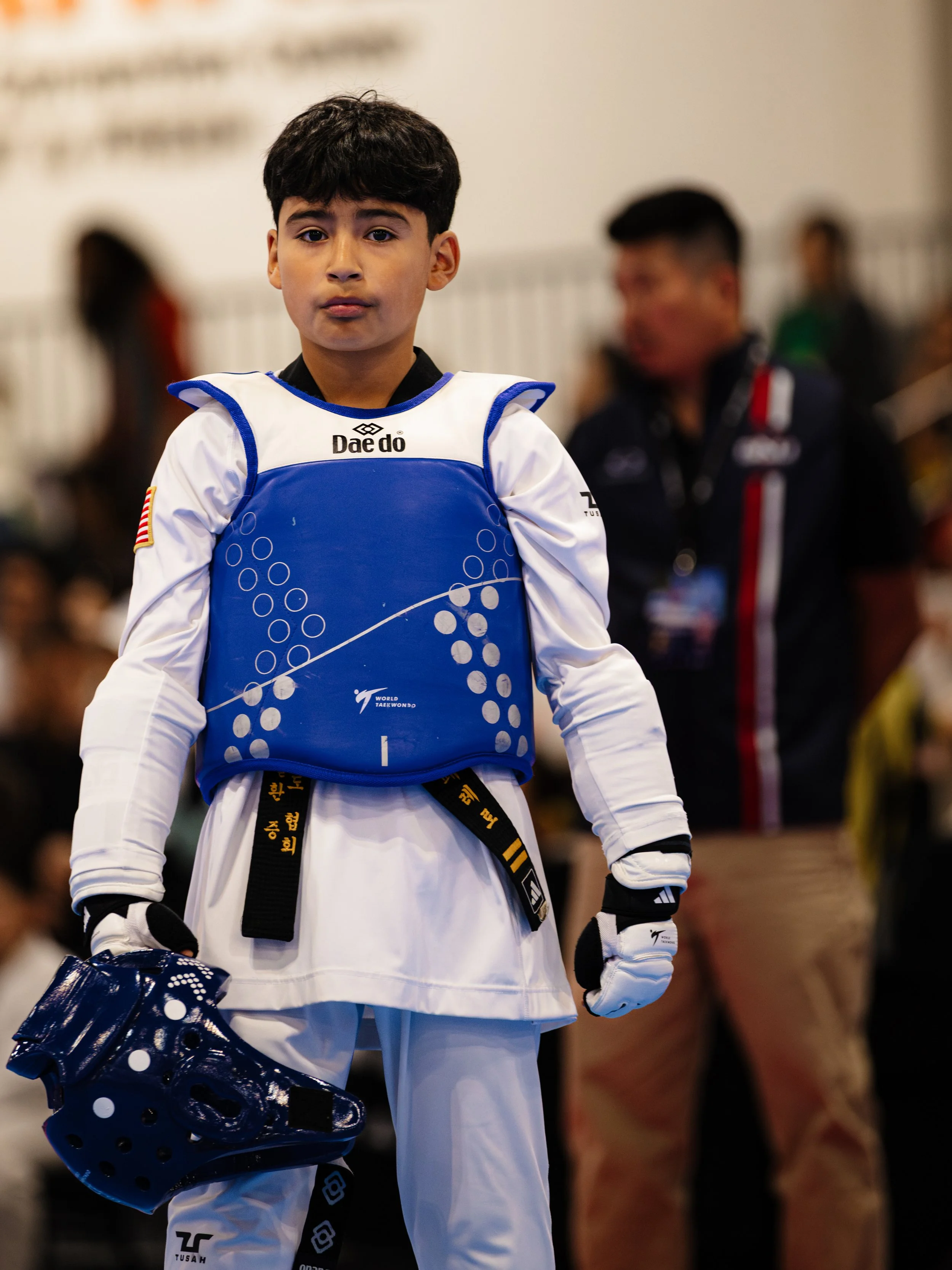Young male taekwondo athlete wearing a white uniform, blue chest protector, black belt, and gloves, holding a helmet, standing in a sports arena.