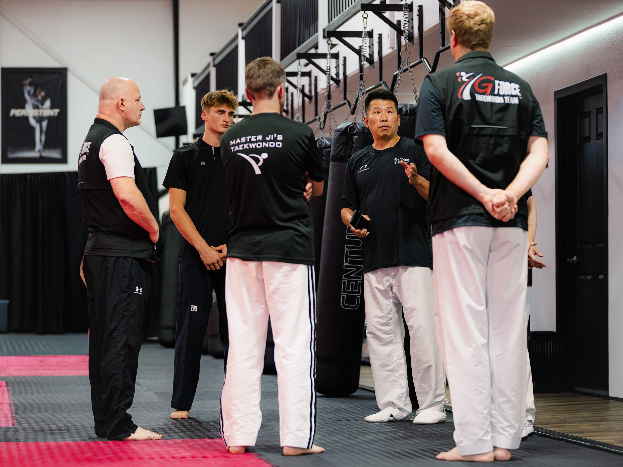A martial arts instructor and five students in a taekwondo gym, standing on black and red mats, listening to the instructor's instructions.
