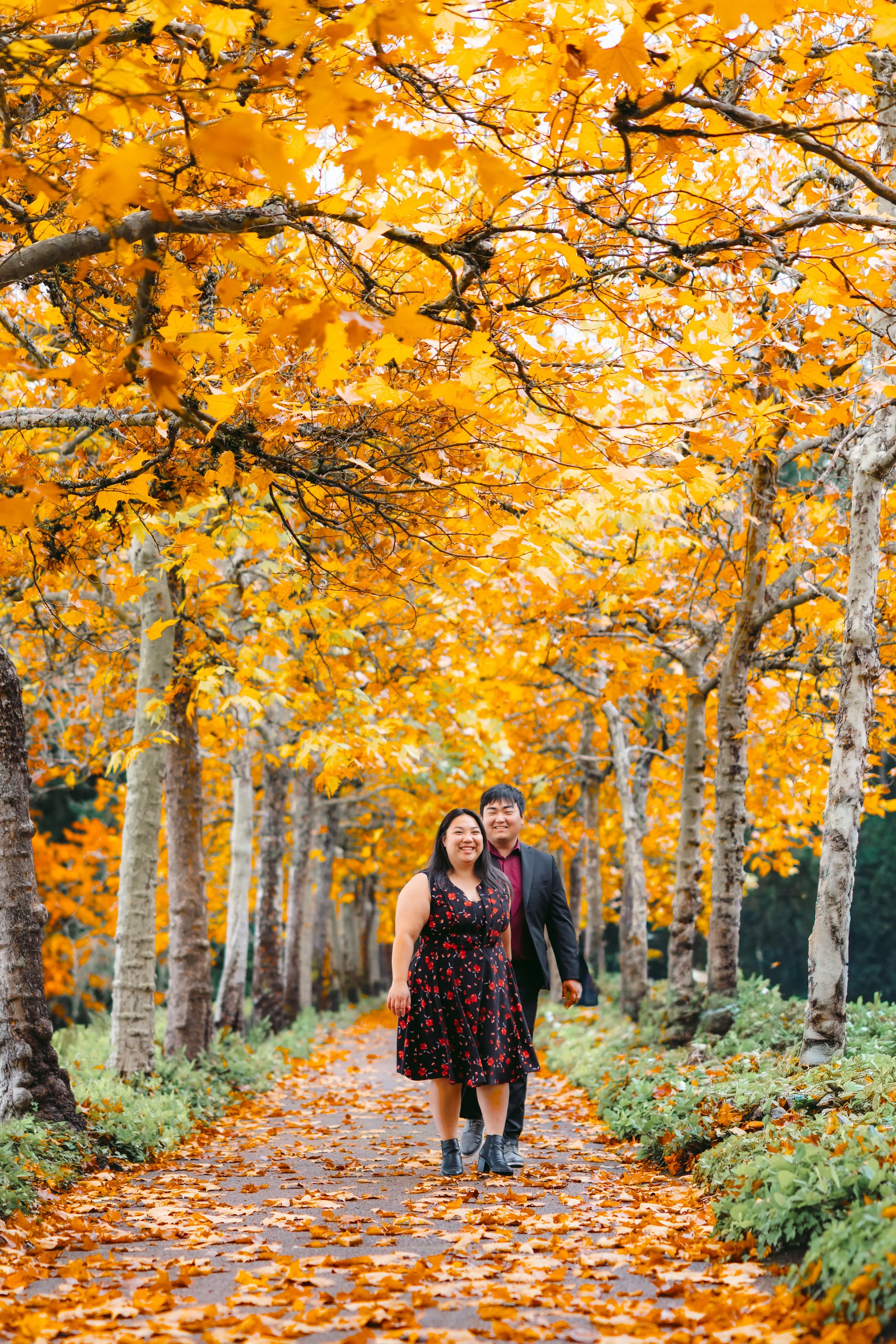 A smiling couple walking arm in arm on a leaf-covered path surrounded by orange autumn trees.