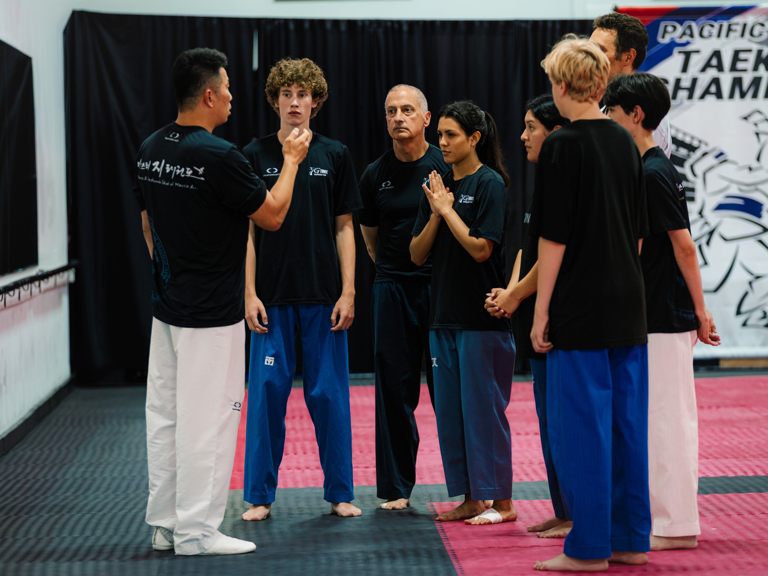 Group of people participating in a martial arts class, listening to an instructor in a gym with a martial arts banner in the background.