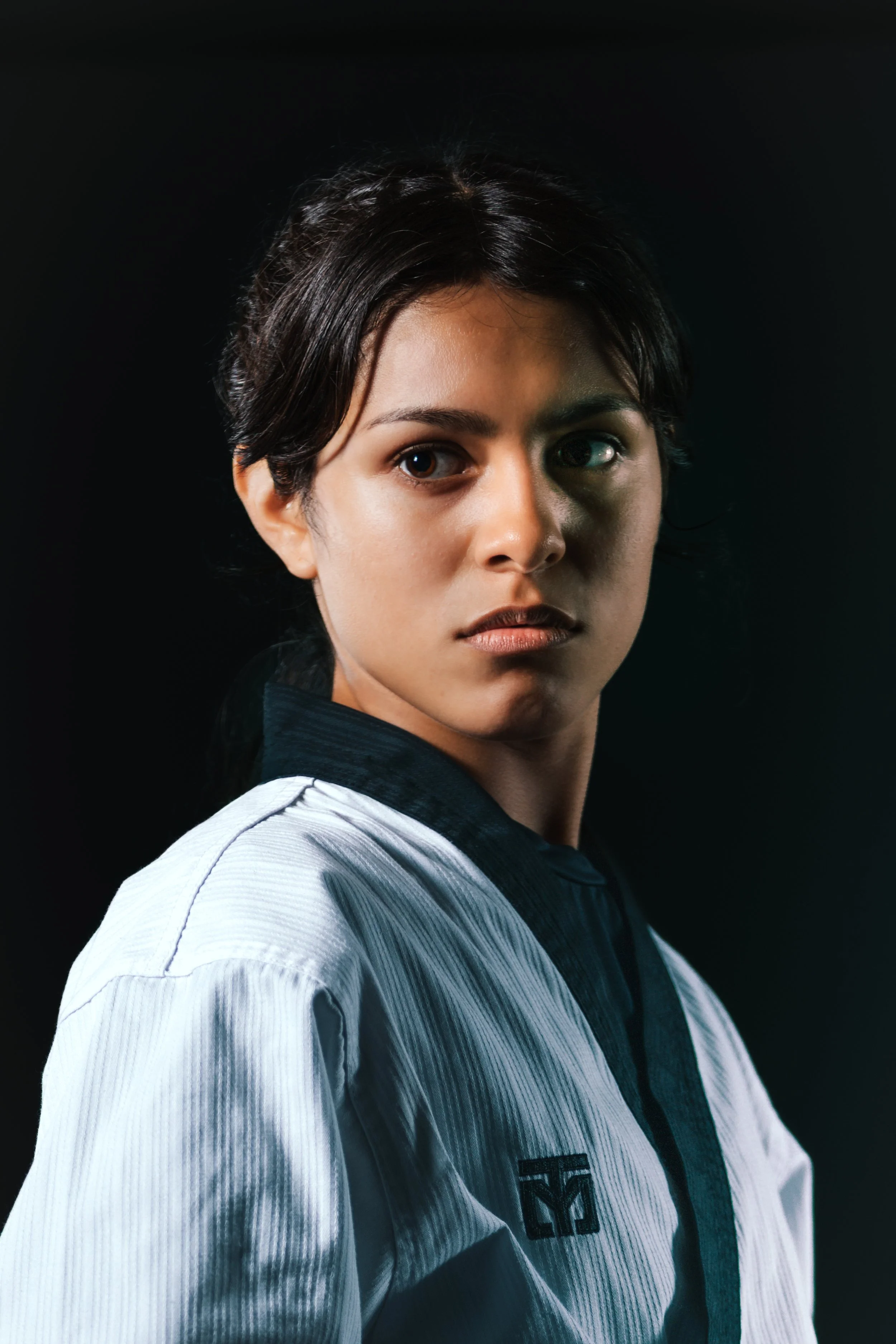 Portrait of a young woman with short dark hair, wearing a white and black martial arts gi, looking over her shoulder against a dark background.