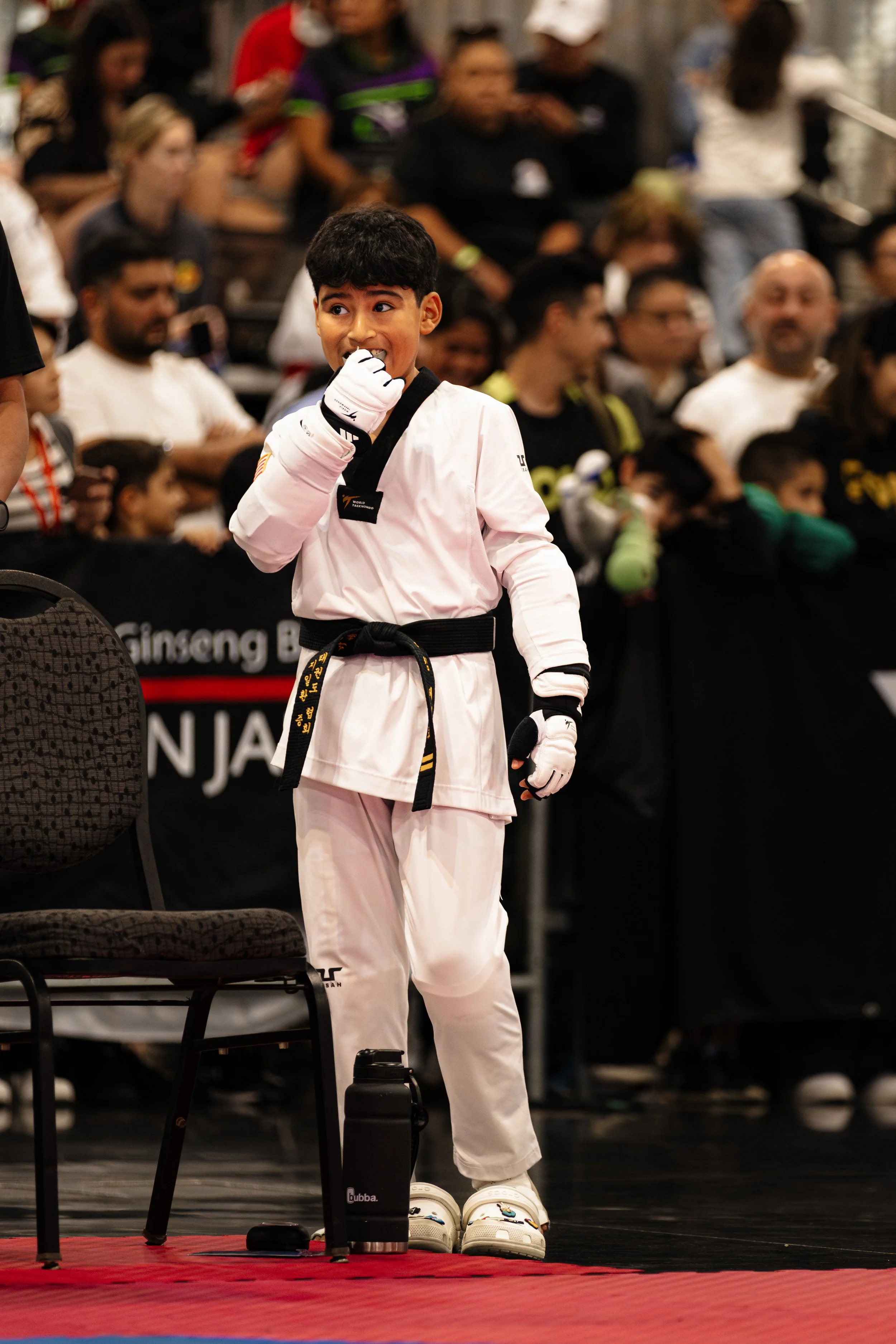 A young male martial artist in a white karate uniform with a black belt stands on a competition mat, adjusting his hand glove, with a group of spectators behind him watching.