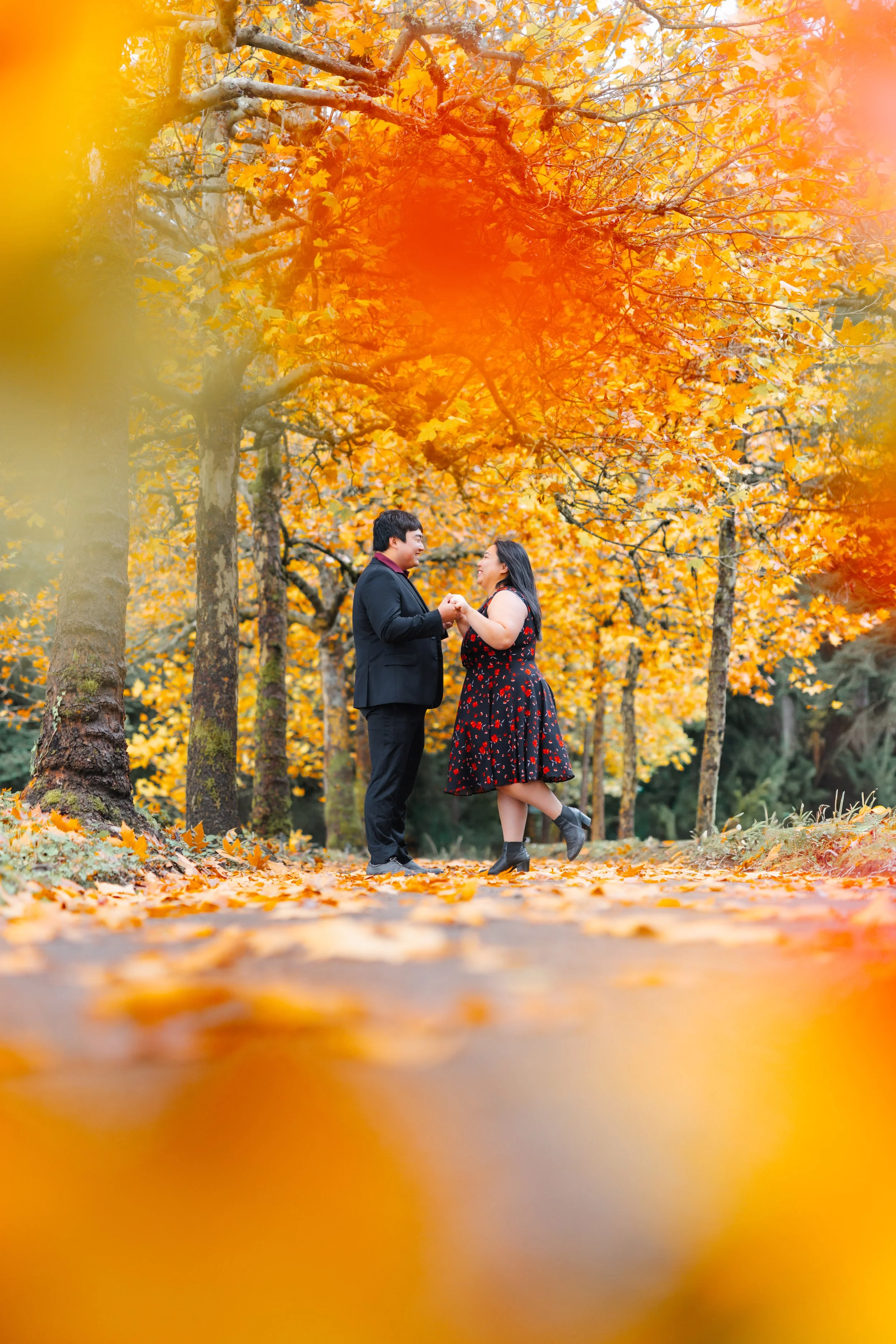 A couple holding hands and dancing in a forest with vibrant orange and yellow autumn leaves.