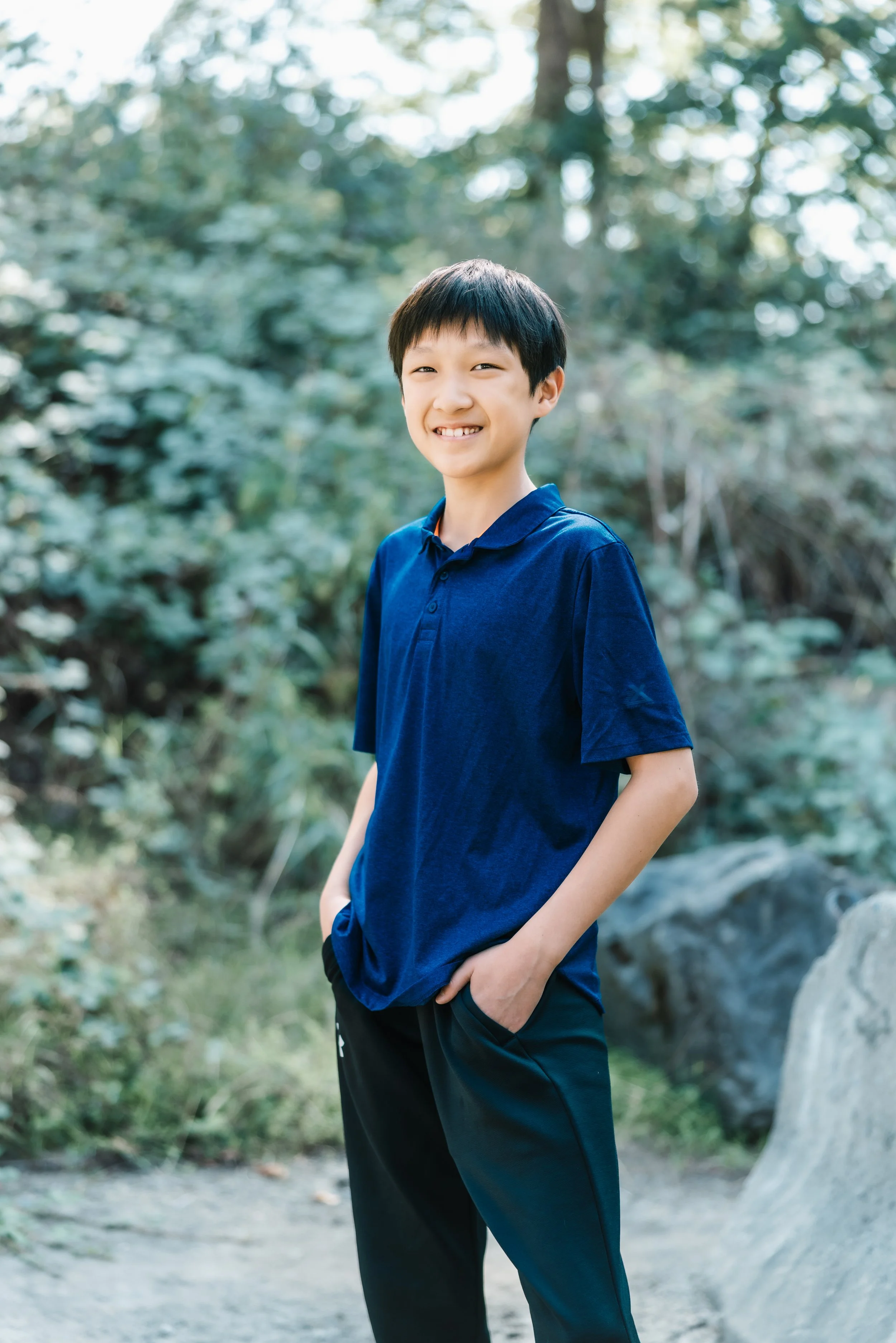 A young boy with dark hair, smiling, wearing a blue polo shirt and black pants, standing outdoors in a lush, green, wooded area.