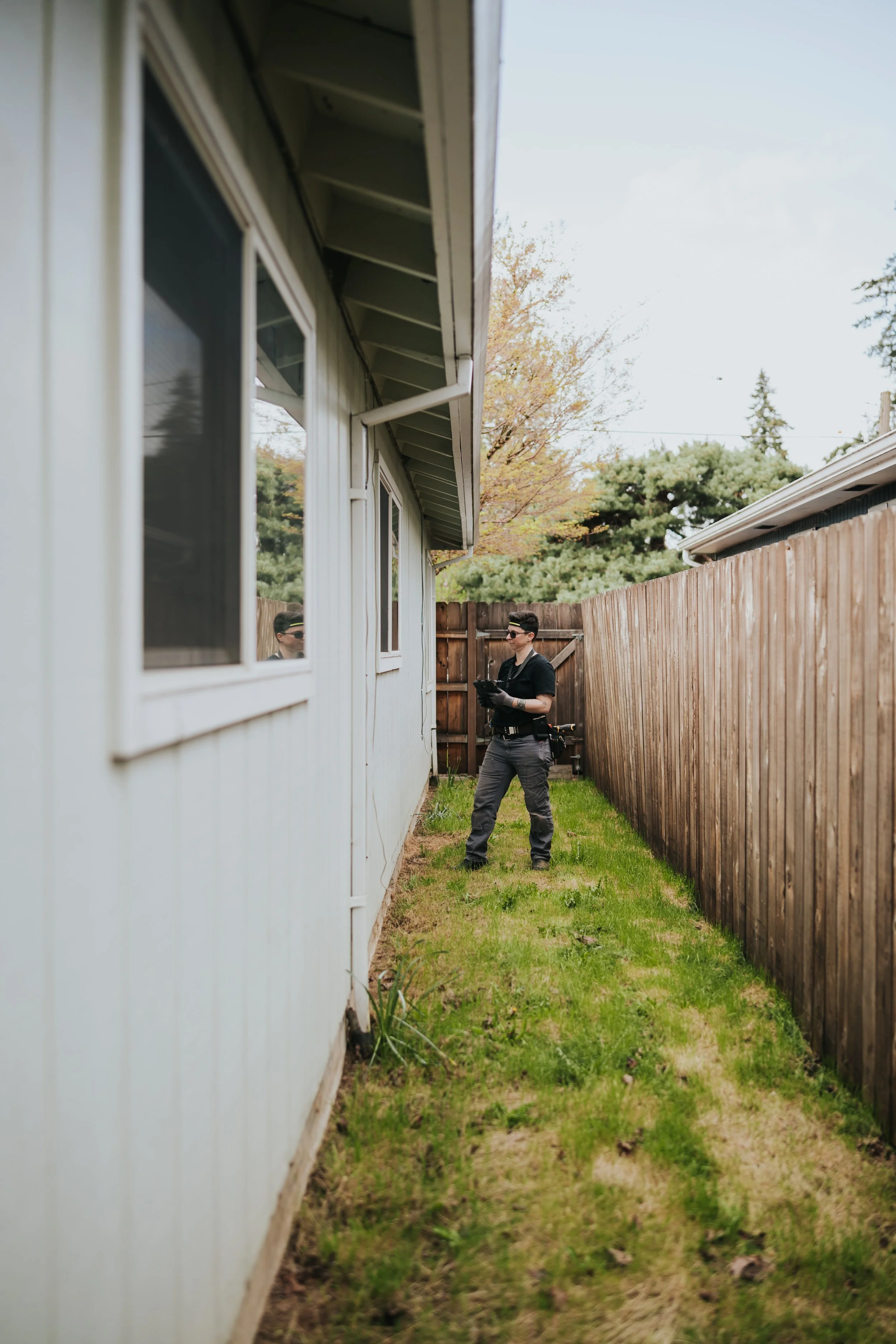 Marissa Longpeak, inspector is wearing protective gear and sunglasses standing in a narrow grassy backyard, holding a tablet, beside a white house with a wooden fence behind.