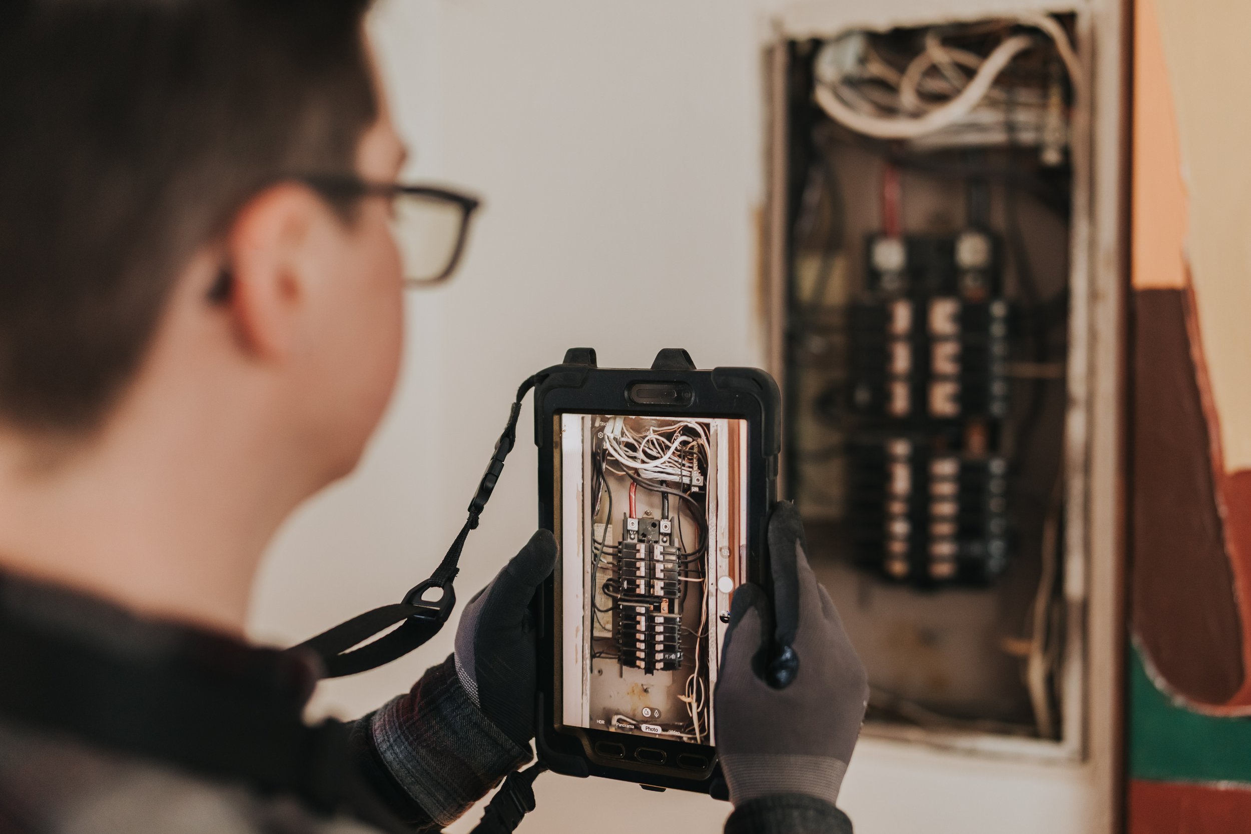Marissa Longpeak, inspector in gloves taking a close-up photo of an open electrical panel with a smartphone.