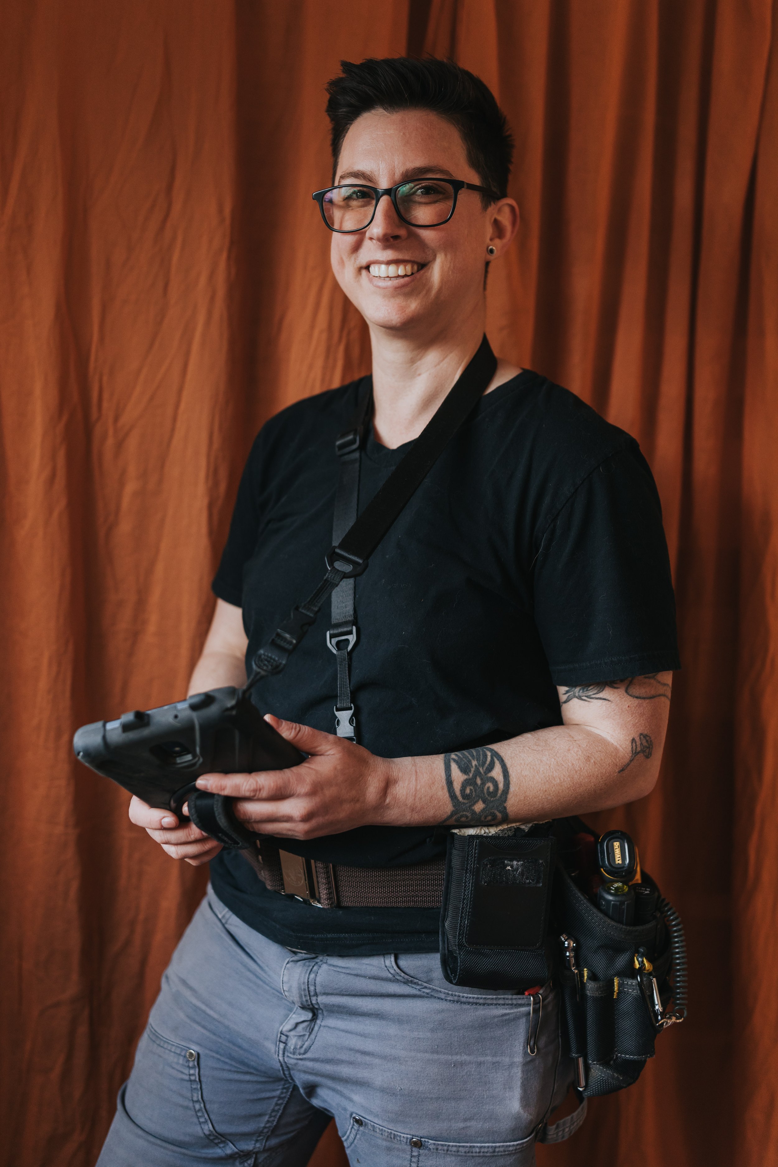 Marissa Longpeak, inspector is smiling, holding a tablet, standing in front of a brown curtain, wearing a black shirt and utility belt with tools.