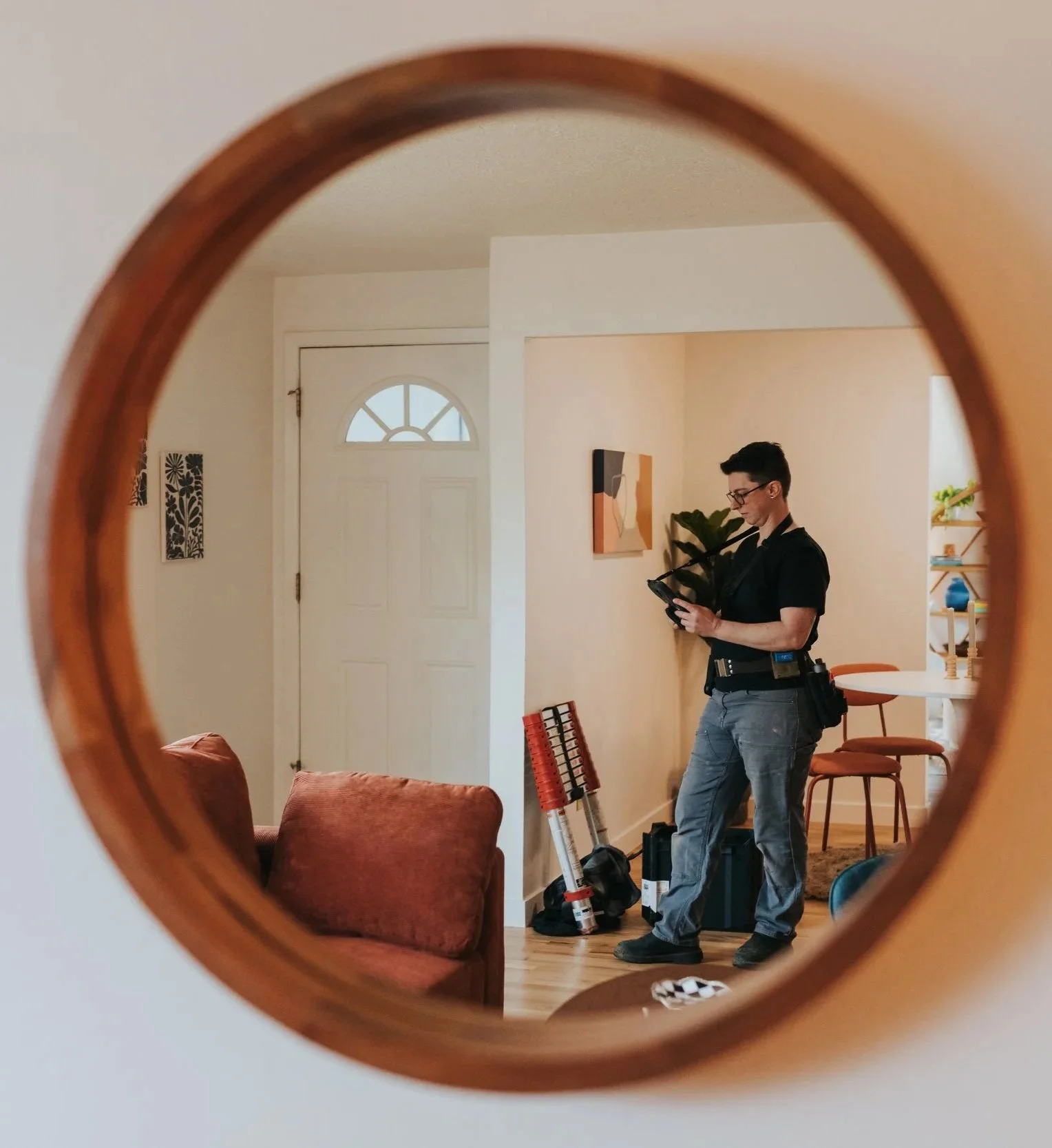 Marissa Longpeak, inspector is wearing a black shirt, jeans, and glasses, standing inside a living room, seen through a circular window, looking at his phone with various tools and equipment nearby.