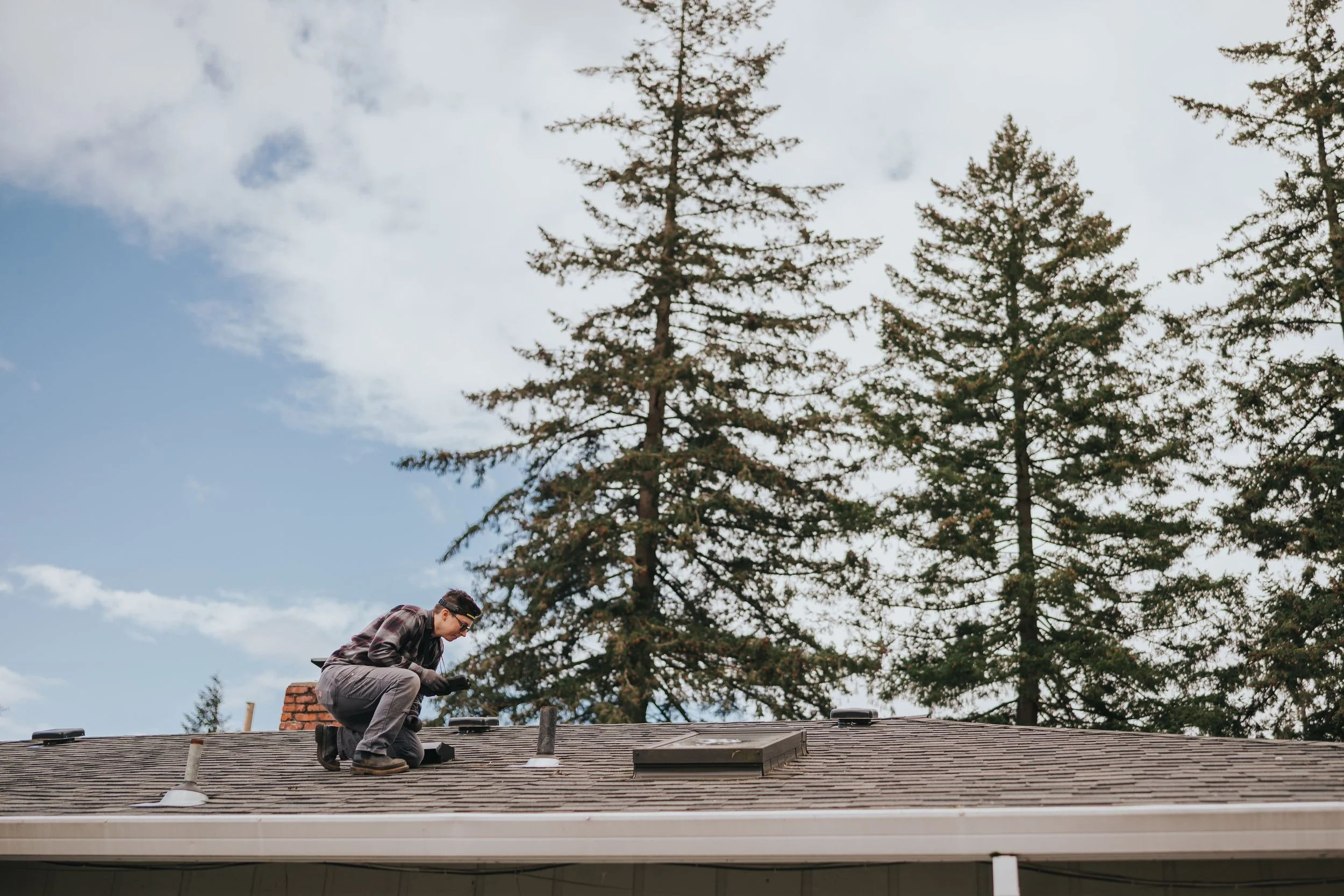 Marissa Longpeak, inspector is kneeling on a house roof performing maintenance or inspection, with trees and a partly cloudy sky in the background.