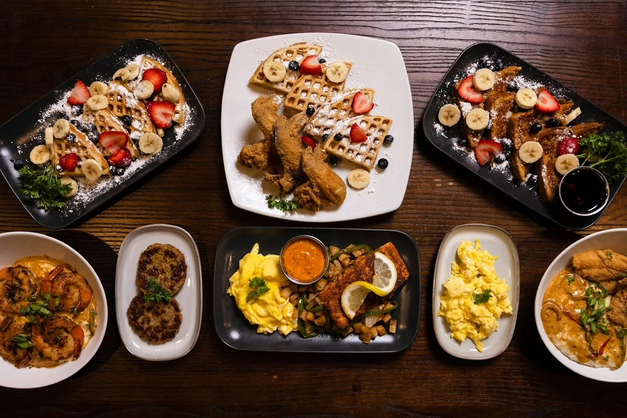 Assorted breakfast dishes on a wooden table, including waffles with strawberries and bananas, fried chicken with berries, scrambled eggs, a fish fillet with lemon, and shrimp in sauce.