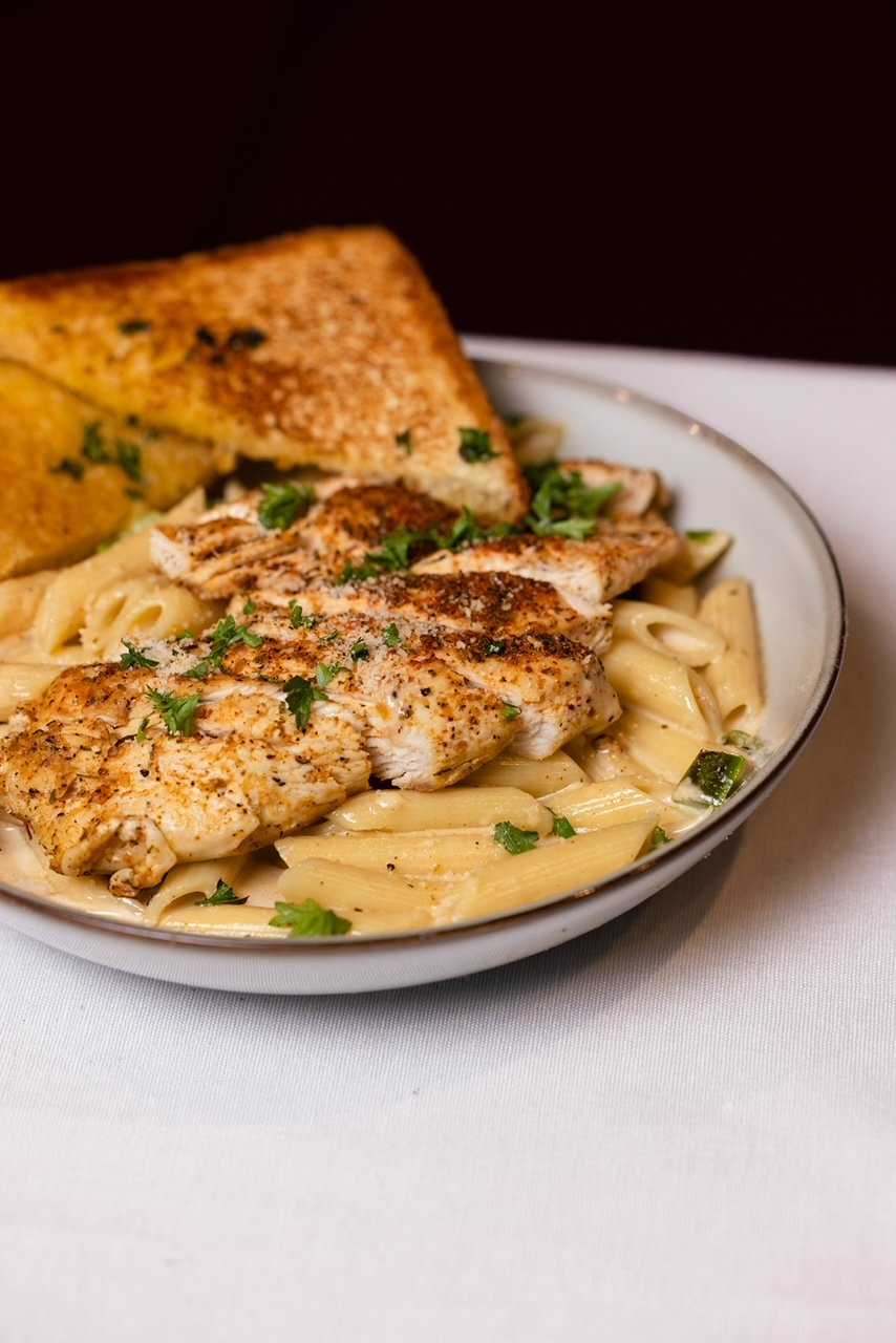 Bowl of creamy pasta with grilled chicken, garnished with parsley, served with garlic bread.
