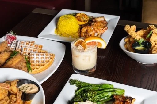 Assorted Asian dishes on a dark wooden table, including fried chicken, rice, grilled vegetables, spring rolls, and a cocktail with a lemon slice.