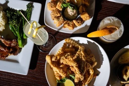 Assorted dishes and drinks on a dark wooden table, including fried chicken, broccoli with steak, and cocktails with lemon garnishes.