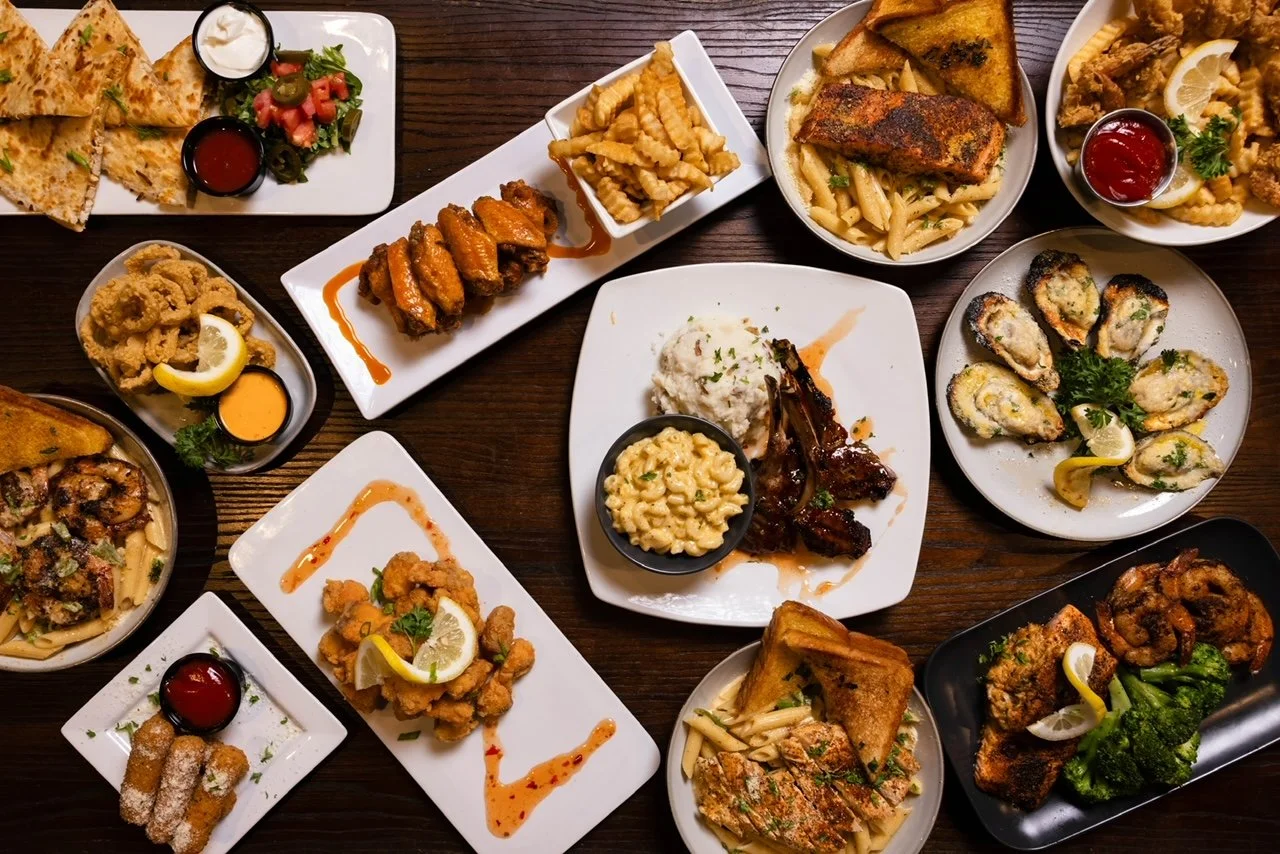 Various plates of Indian and Mediterranean food including fried chicken, grilled fish, pasta, stuffed oysters, fries, bread, and vegetables on a wooden table.