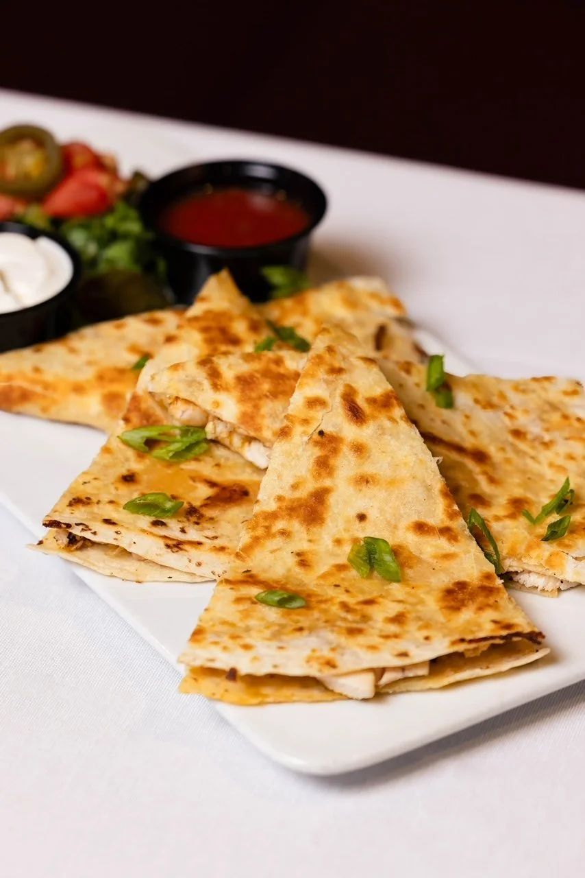 Quesadilla on a white plate with green onions, served with salsa and sour cream in black containers in the background.