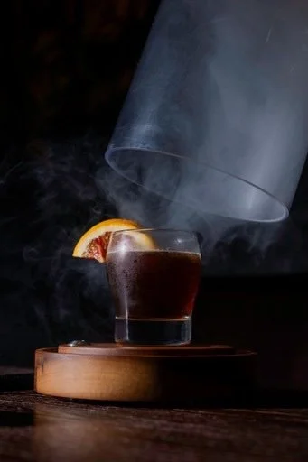 A glass of dark beverage with a lemon wedge, sitting on a wooden coaster, with smoke rising around it, against a dark background.