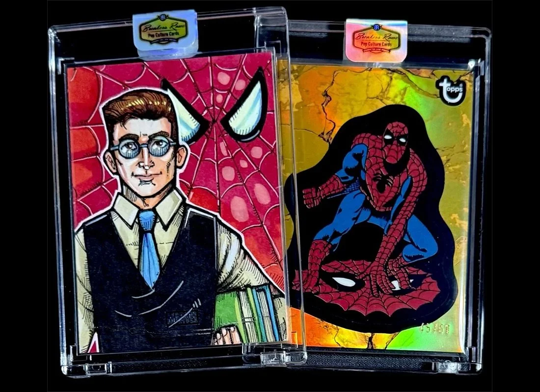 Two collectible trading cards inside a clear plastic case. The card on the left features a comic-style illustration of a man with glasses, dressed in a white shirt, blue tie, and black vest, with a comics background of a spider web. The card on the r