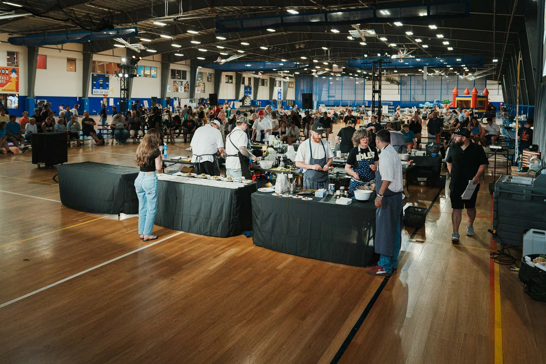 View of an indoor event in a gymnasium with people sitting and standing. There is a food table with chefs preparing dishes and a crowd seated in the background. The gym has a high ceiling with overhead lights and a wooden floor.