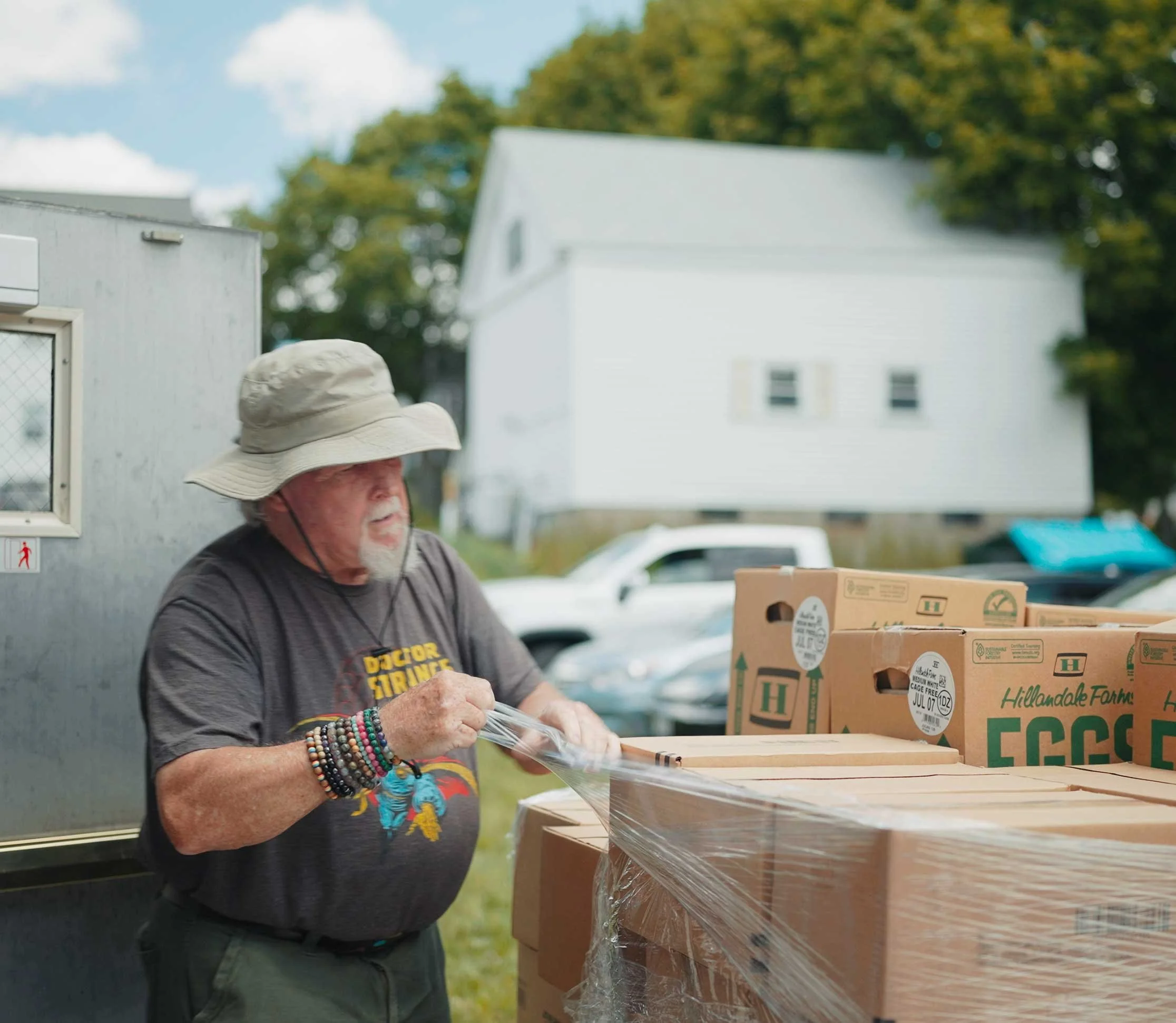 An elderly man with a large hat and multiple colorful bracelet stacks on his wrist is wrapping a plastic sheet around boxes of eggs at an outdoor farm stand.