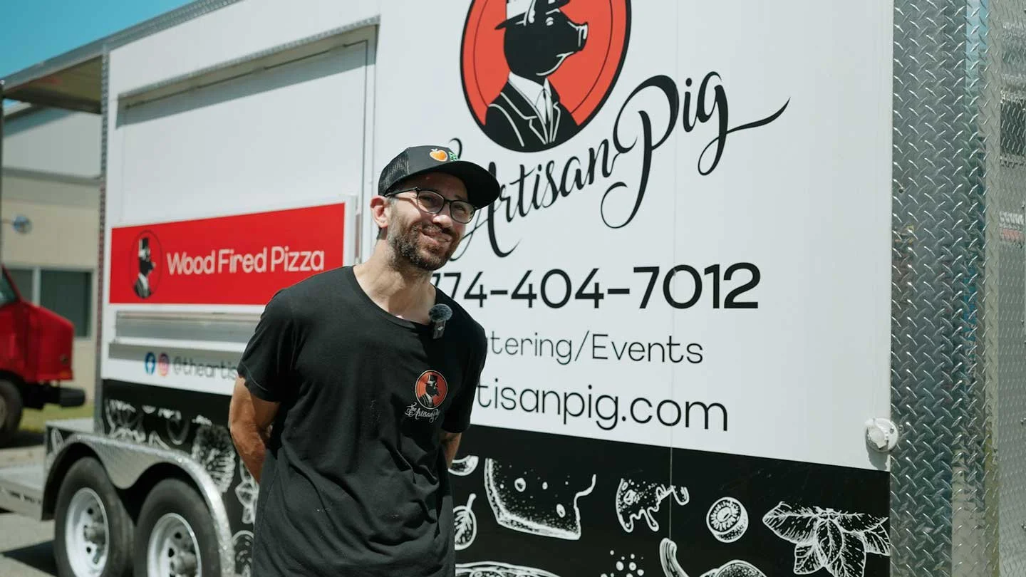 A man wearing a black t-shirt, glasses, and a black cap stands in front of a large white food truck with a red and black logo of a pig in a suit. The truck has the text "Artisan Pig," a phone number, and a website on its side. There are additional illustrations at the bottom of the truck, including slices of bread, cheese, and vegetables.