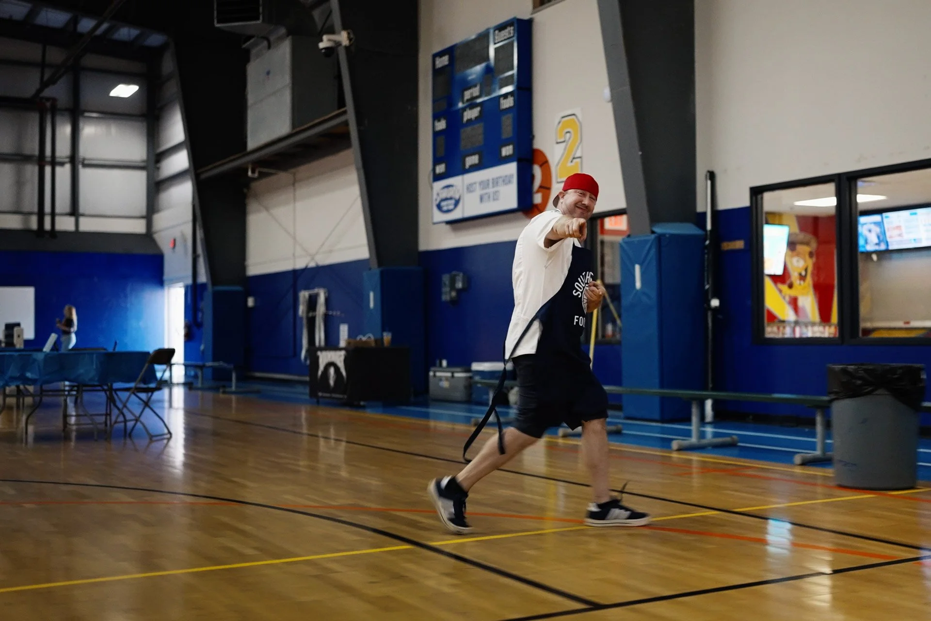 A man wearing a red cap, black shorts, and a black t-shirt with white text inside a gymnasium, smiling and pointing towards the camera while holding a resistance band. There are tables and chairs to the side, a scoreboard above, and a woman in the background near the wall.